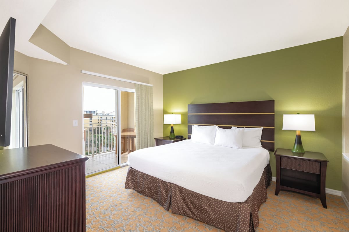 A hotel room with a large bed, two bedside tables with lamps, a TV, and a balcony with a view of buildings.