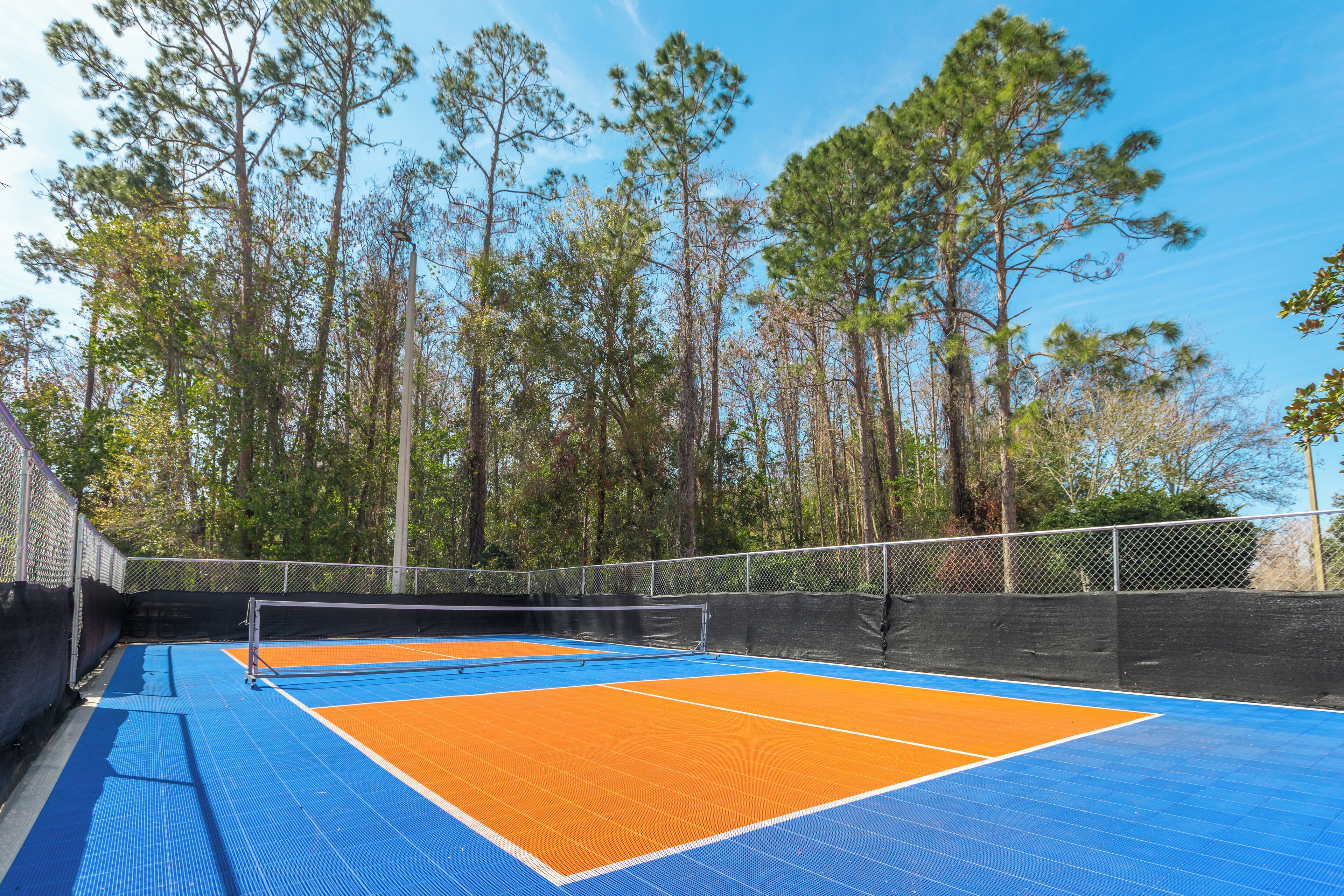 An outdoor tennis court with blue and orange surfaces, surrounded by tall trees.
