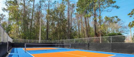 An outdoor tennis court with blue and orange surfaces, surrounded by tall trees.