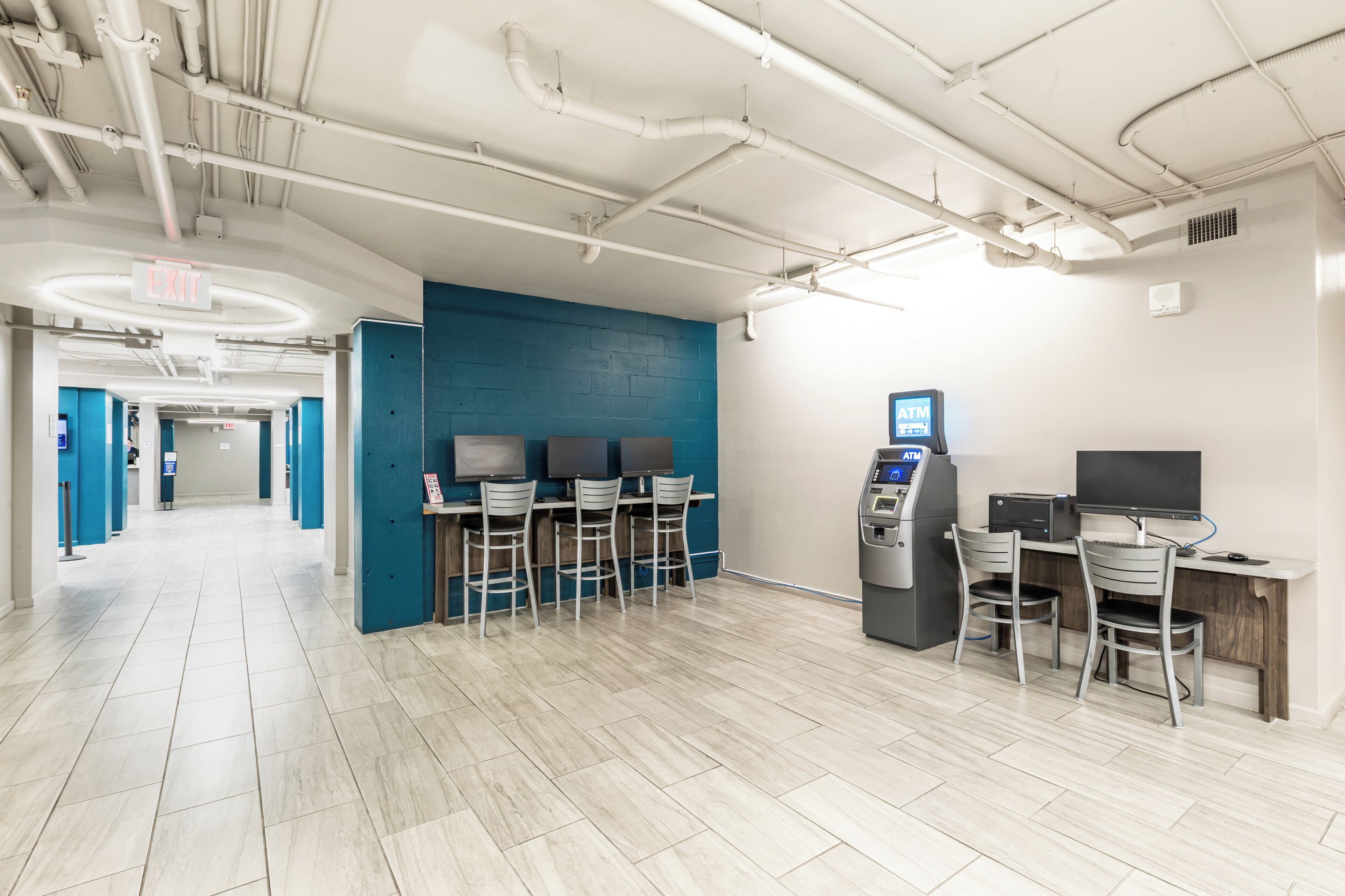 A modern indoor space with a blue accent wall, a row of bar-style seating, an ATM, and a computer station.