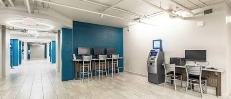 A modern indoor space with a blue accent wall, a row of bar-style seating, an ATM, and a computer station.