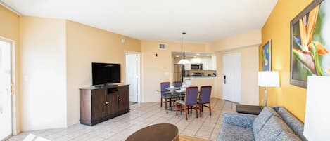 A living room with a sofa, a television, a dining table with chairs, and a kitchen area in the background.