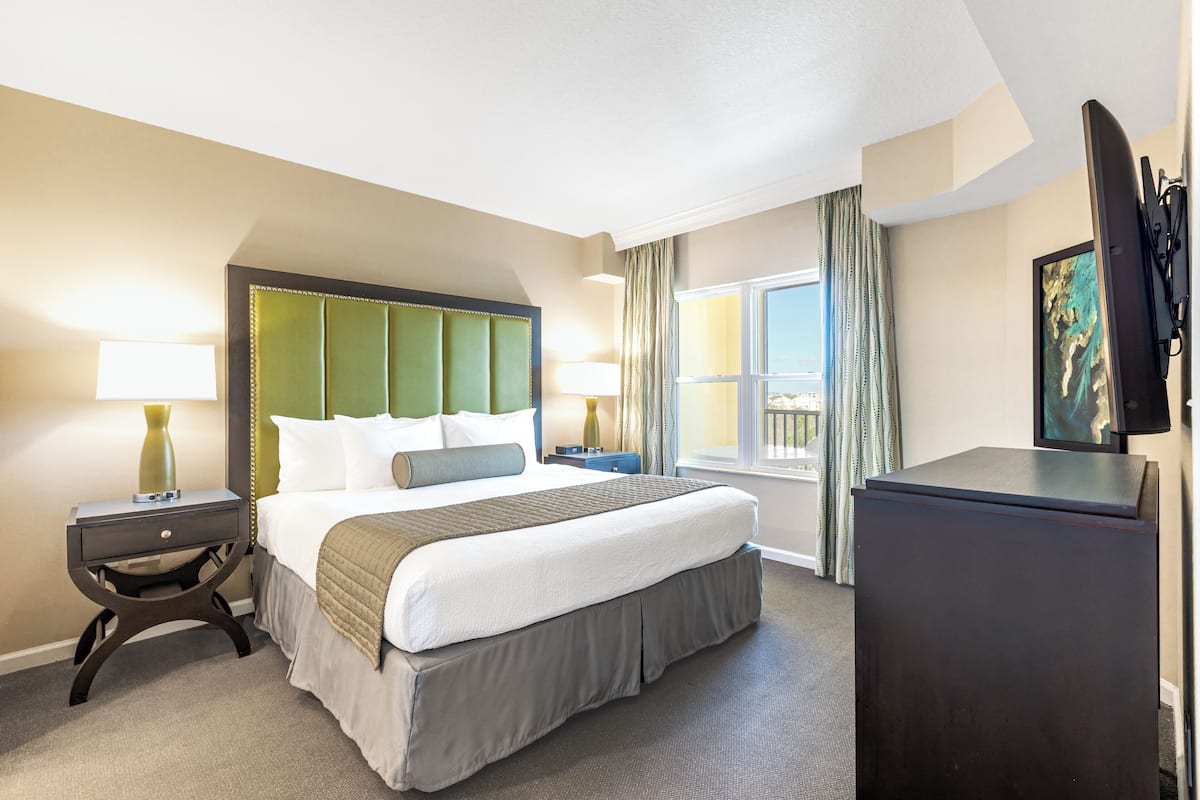 A hotel room with a large bed, a nightstand with a lamp, a dresser, and a window with curtains.