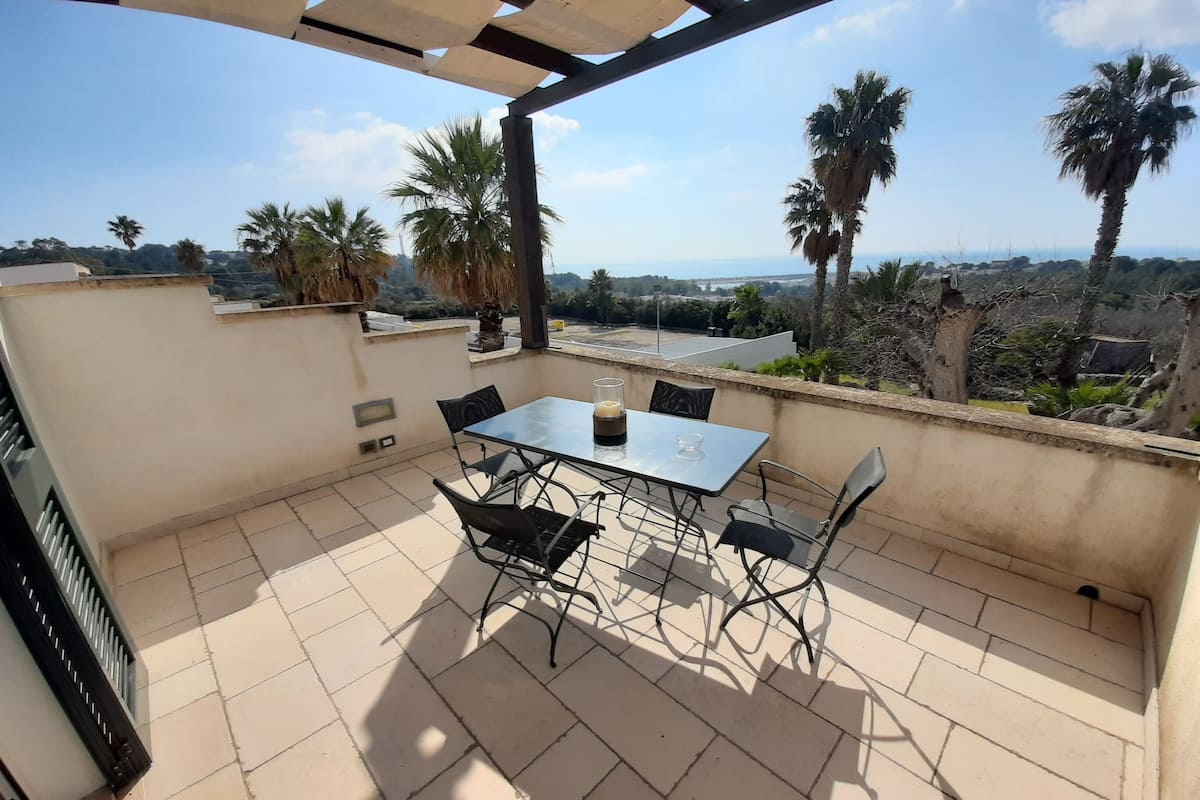 Eine Terrasse im Freien mit Tisch und Stühlen, mit Blick auf eine malerische Landschaft aus Palmen und Meer.