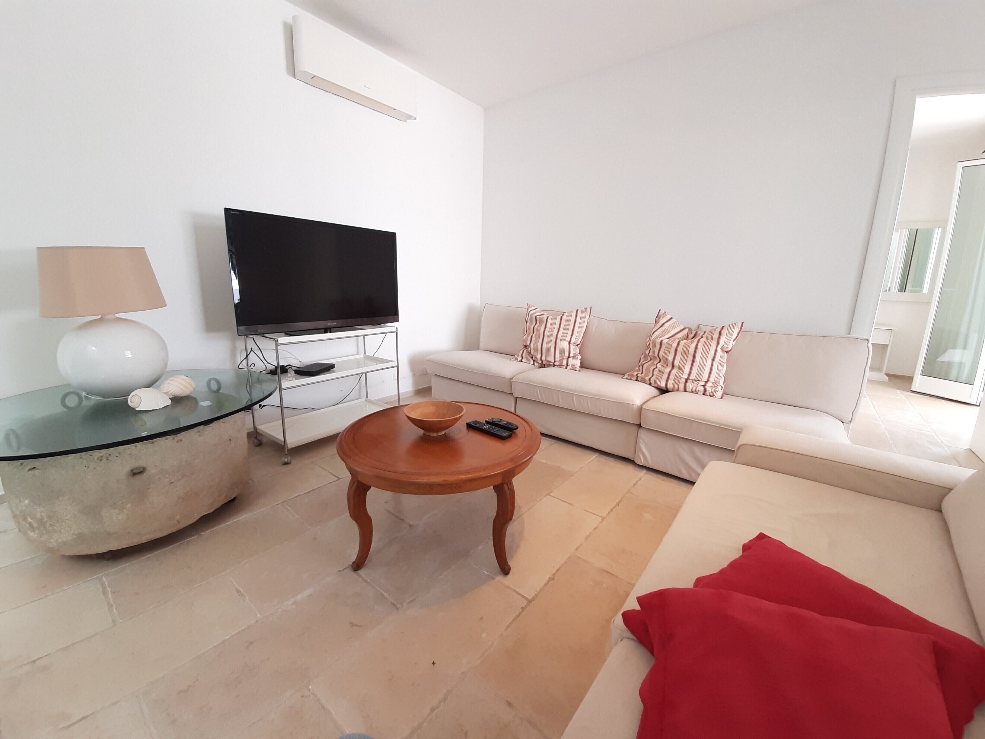 A modern living room with a flat-screen TV, a glass coffee table, a beige sofa, and a red throw blanket.