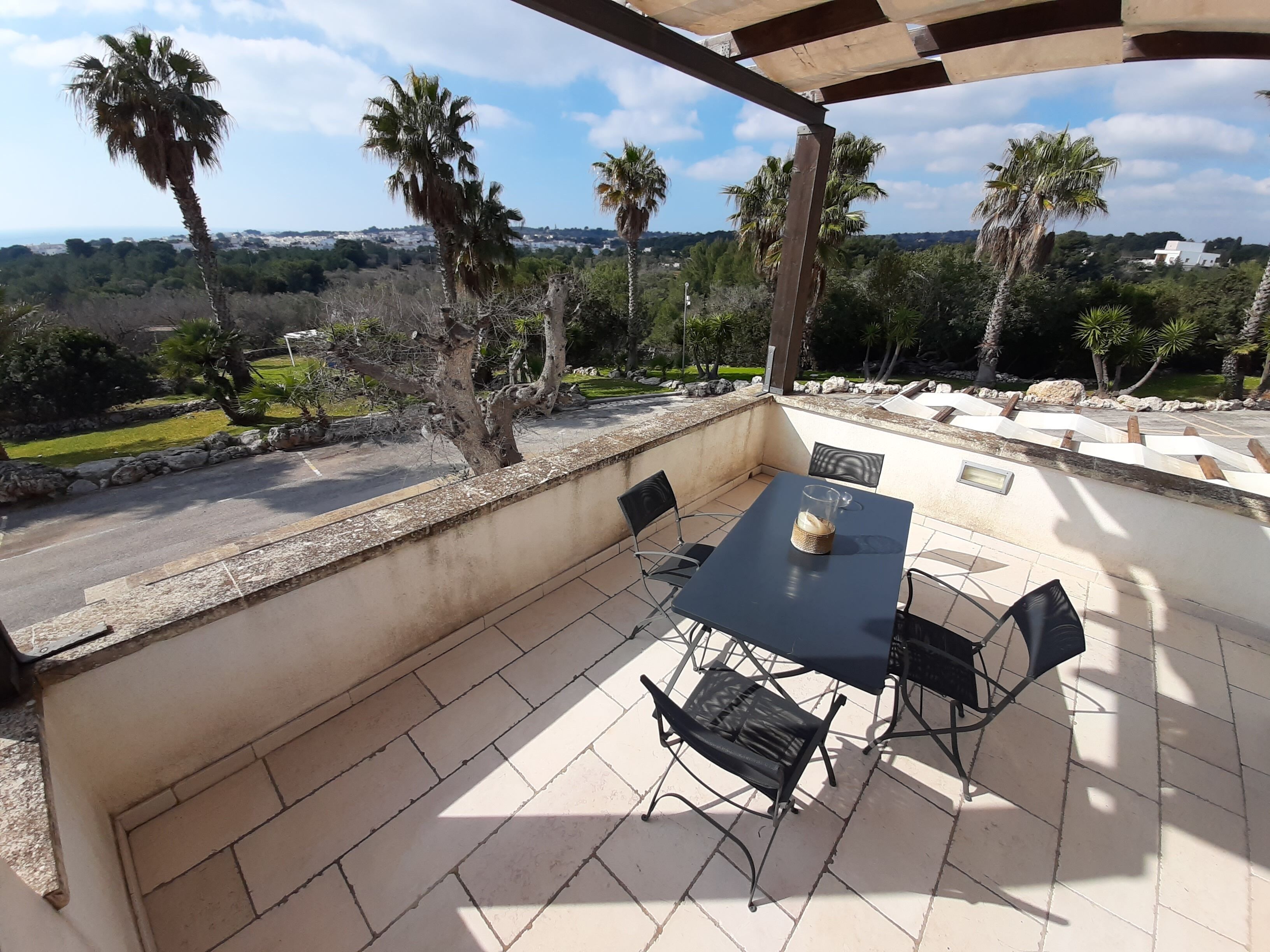 Une terrasse extérieure avec une table et des chaises, offrant une vue panoramique sur un paysage verdoyant, avec des palmiers et des maisons visibles au loin.
