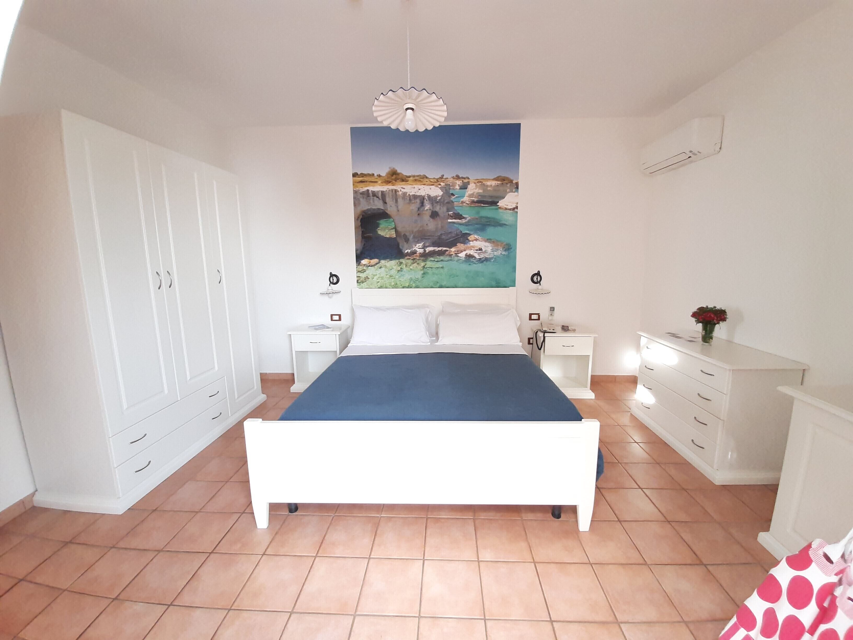Un dormitorio con cama, armario blanco, mesitas de noche y un mural de paisaje costero.