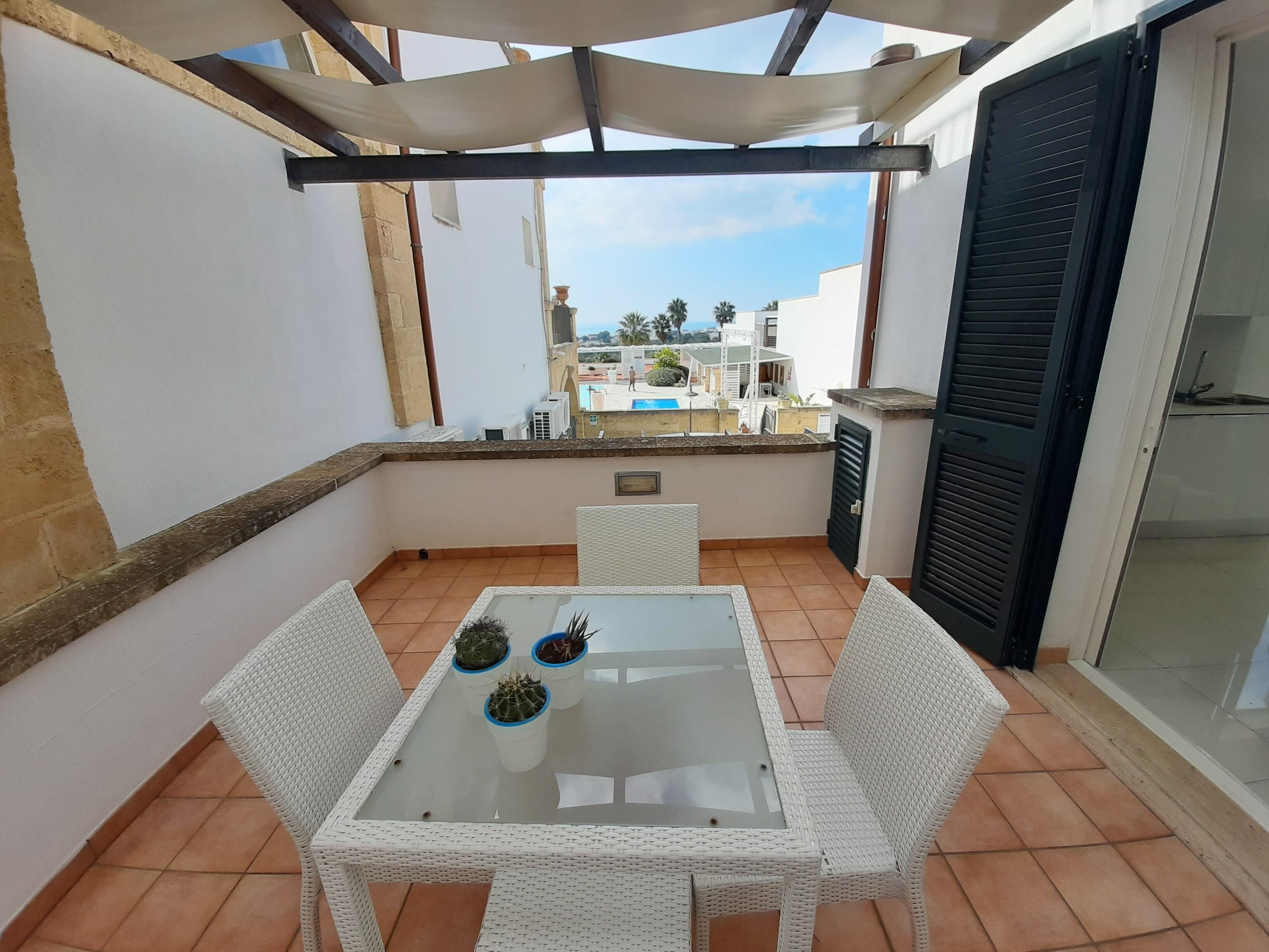 A balcony with a glass table and white wicker chairs, a view of a pool and buildings in the background.