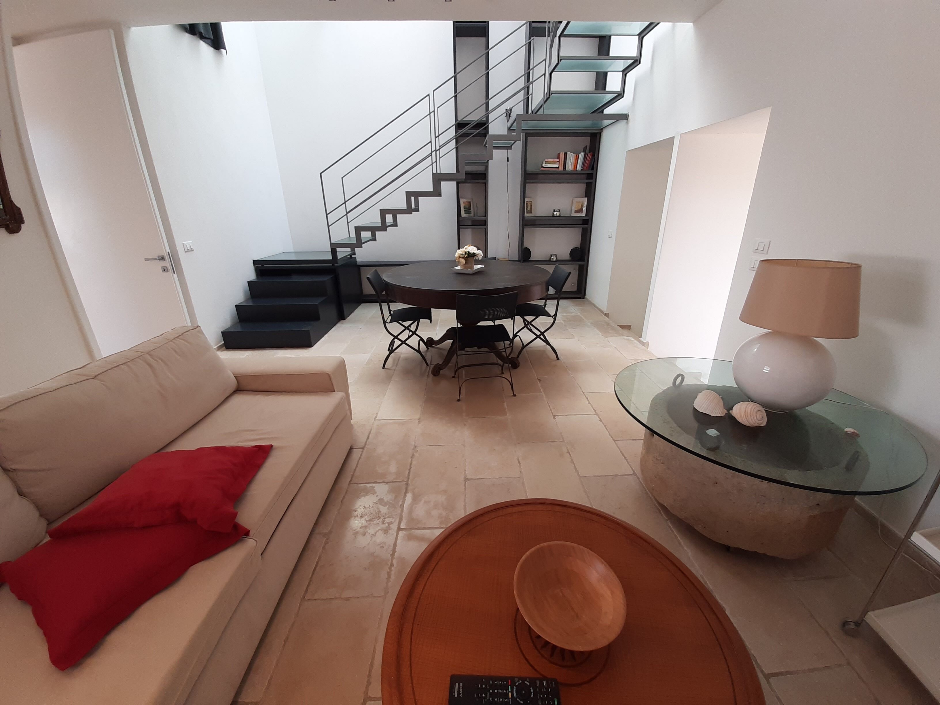 A modern living room with a sofa, a glass coffee table, a dining table with chairs, a staircase, and a bookshelf.