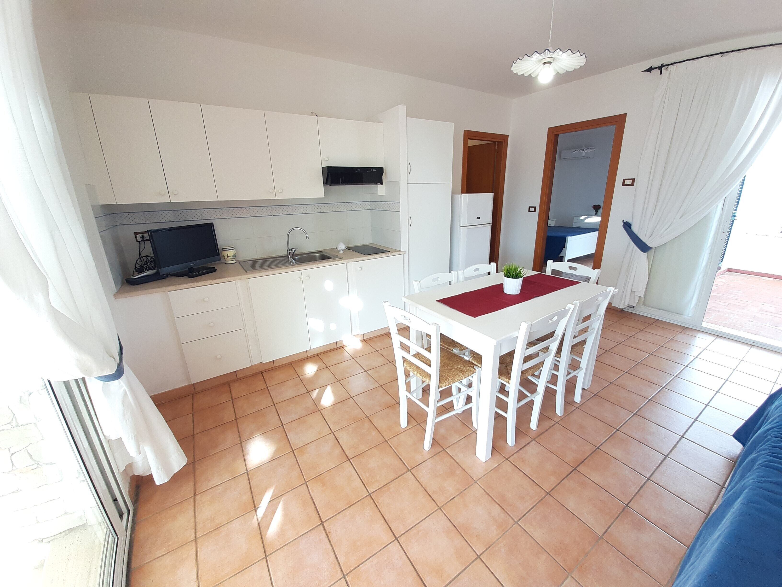Een compacte keuken met witte kastjes, een eettafel met stoelen en een kleine koelkast.