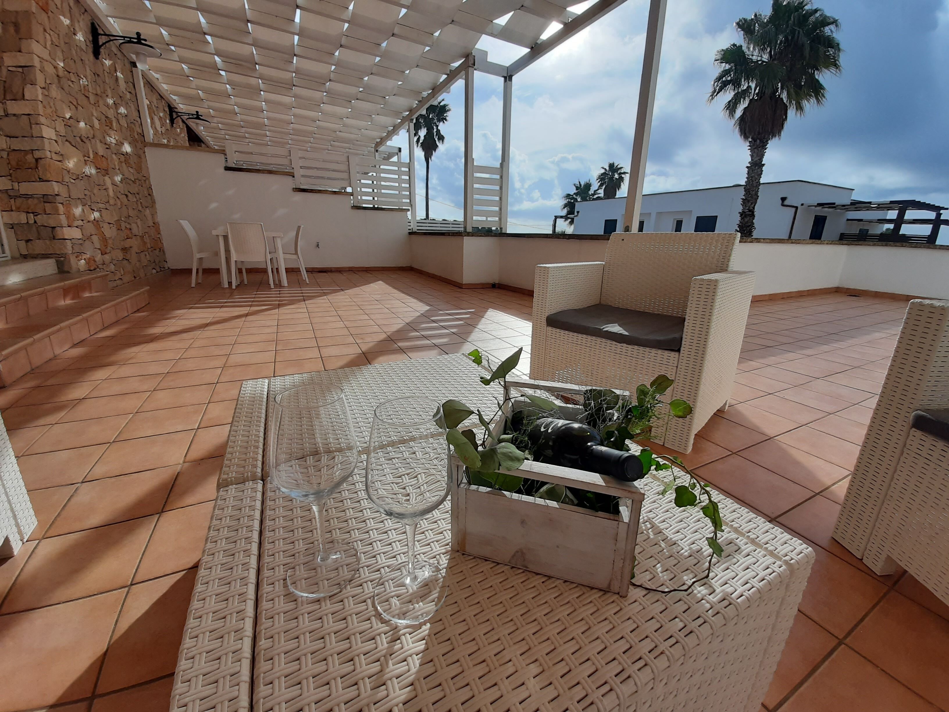 Un patio en la azotea con mobiliario de mimbre, una mesa con copas de vino y una maceta con plantas.