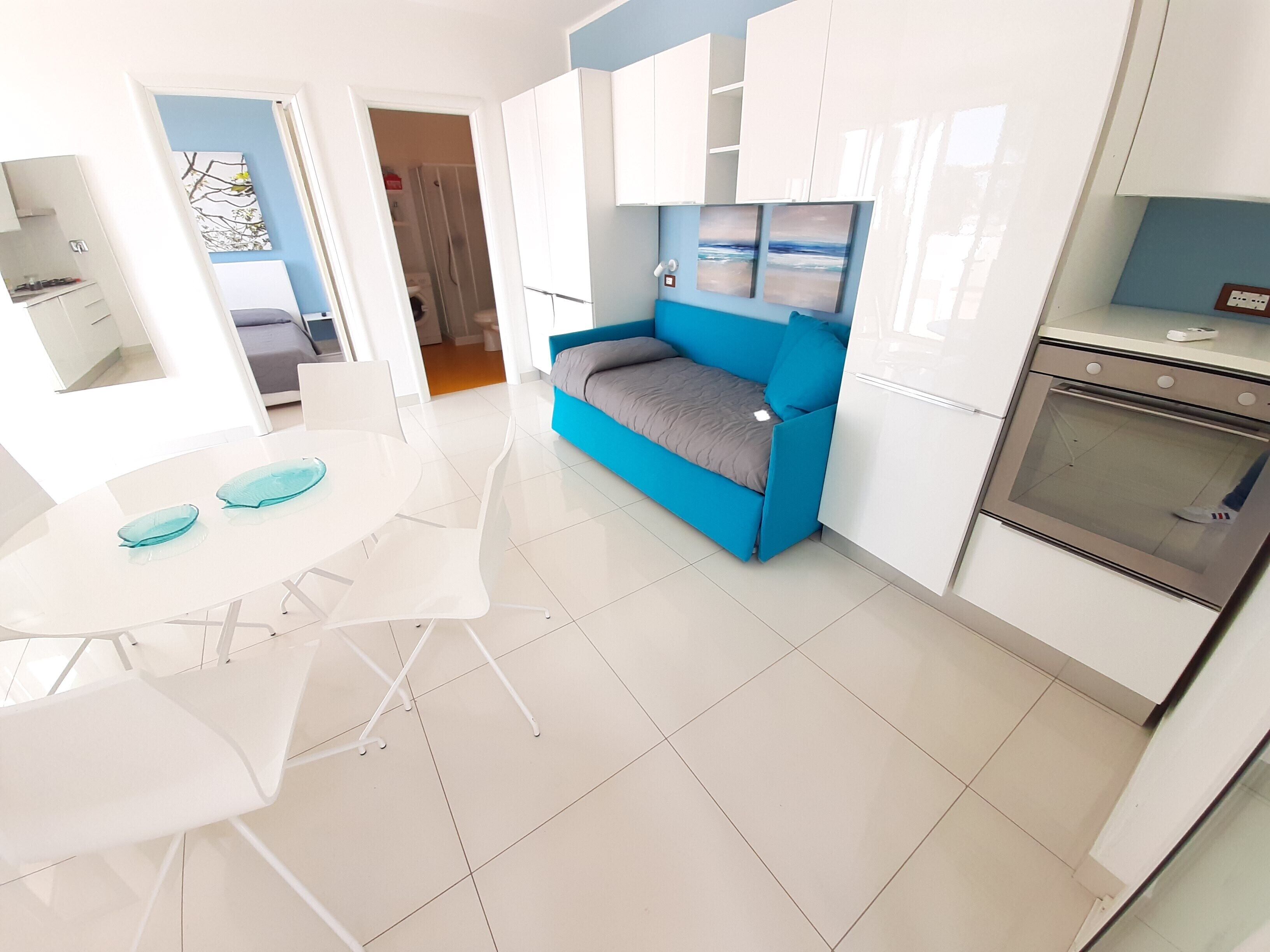 Uma cozinha moderna com armários brancos, um sofá azul e uma área de jantar com mesa e cadeiras brancas.