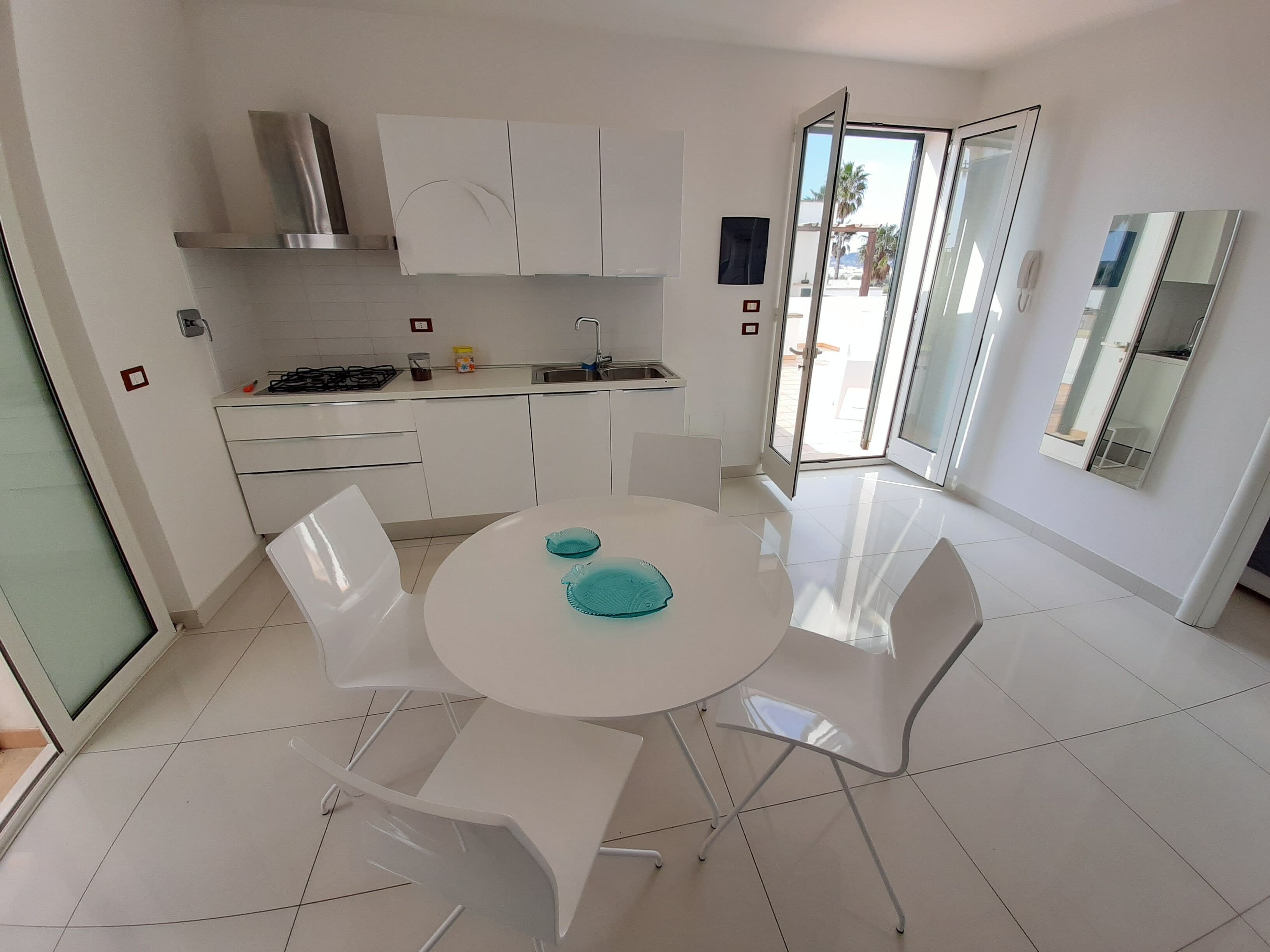 Una cocina moderna con armarios blancos, una estufa a gas y un lavamanos. Hay una zona de comedor con una mesa y sillas blancas, y un ventanal que ofrece vistas al exterior.
