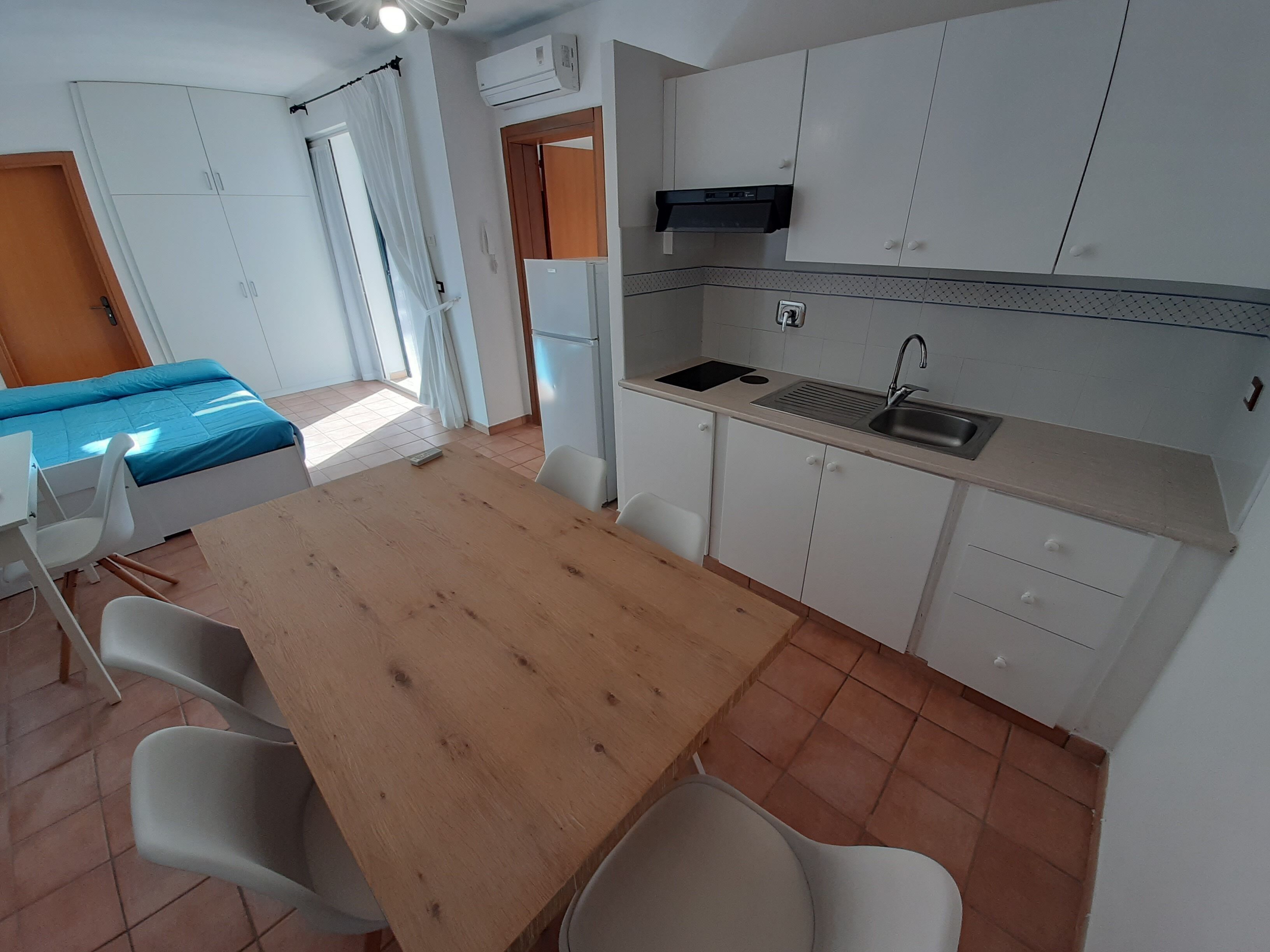 Una cocina compacta con una mesa de madera, sillas blancas, un lavabo y un refrigerador.