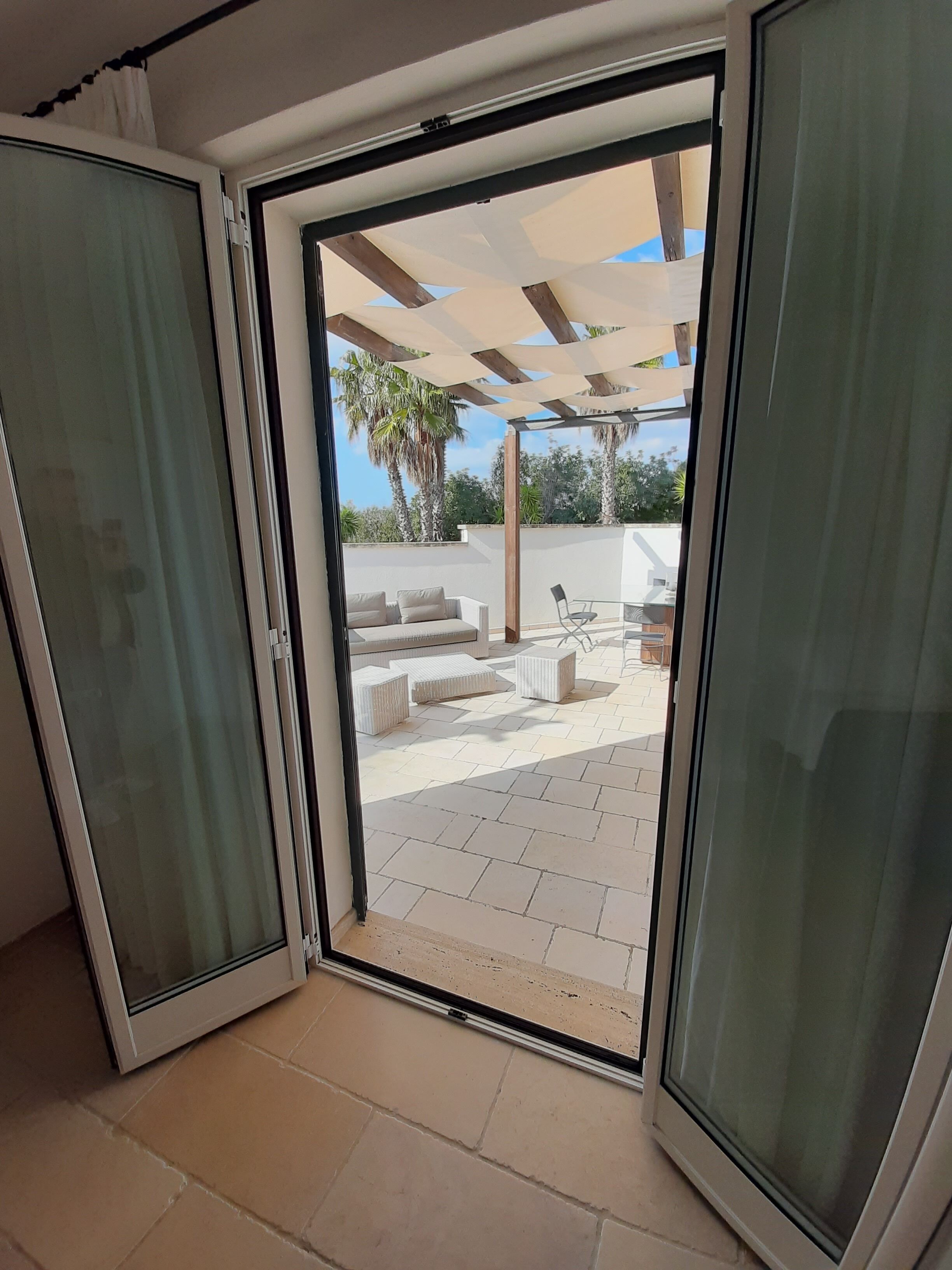 Een schuifdeur die uitkomt op een terras met een pergola, buitenmeubels en een uitzicht op palmbomen.