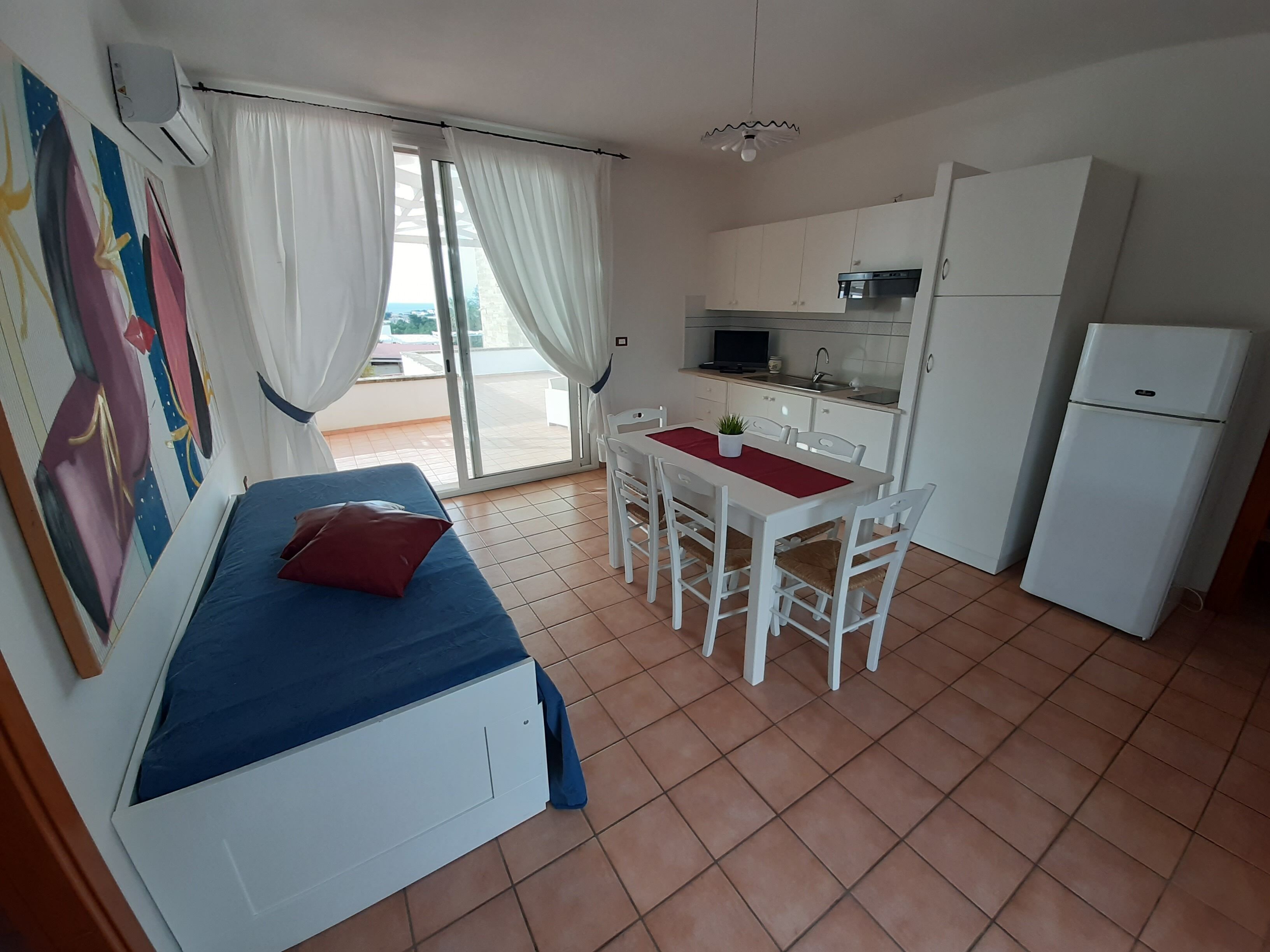 Ein kompakter Raum mit einem Bett, einem Esstisch mit Stühlen, einer Küchenzeile und einem kleinen Kühlschrank.