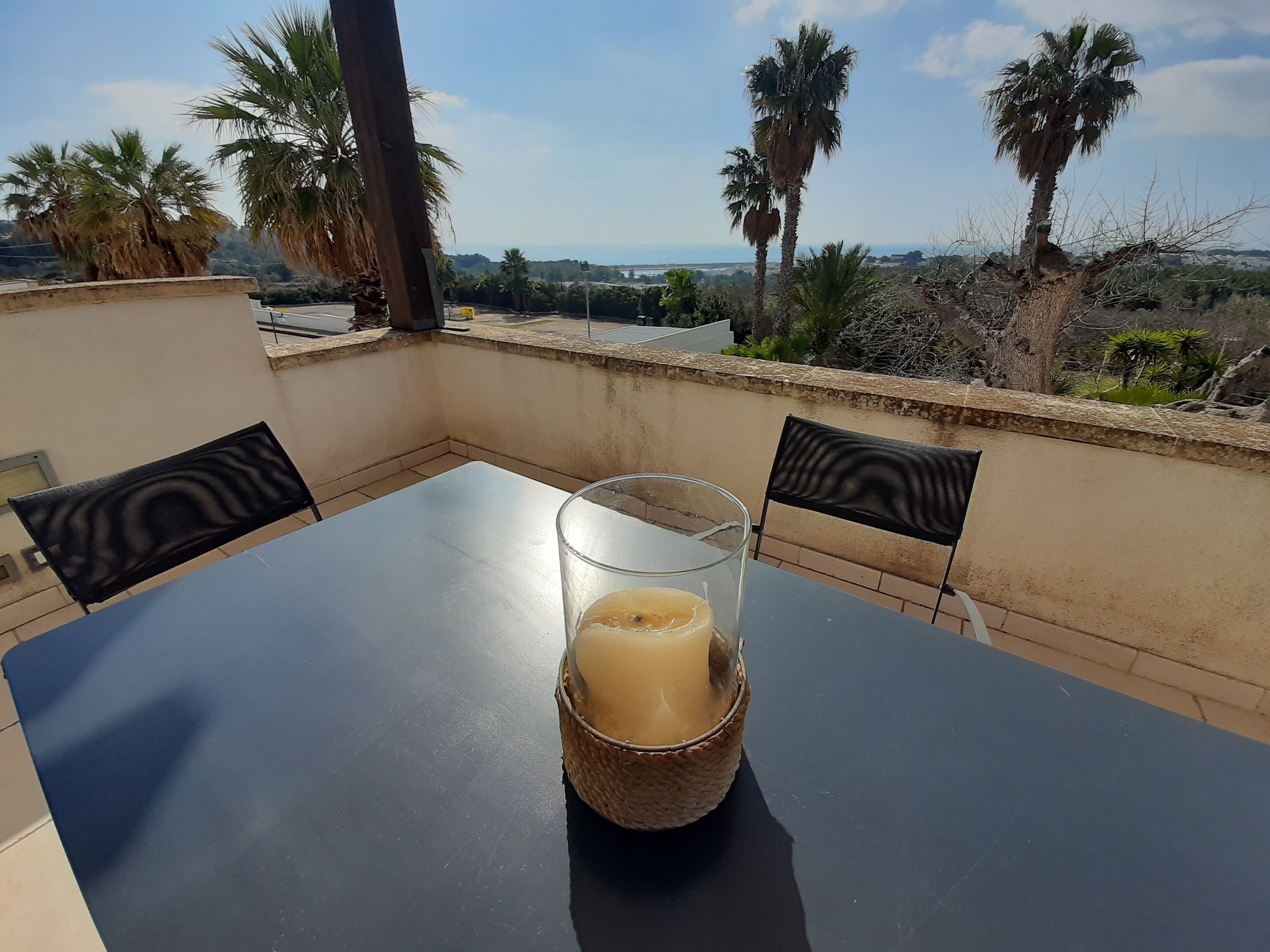 A balcony with a table and chairs, a glass candle holder with a candle, and a view of palm trees and distant landscape.