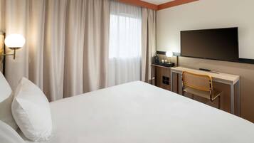 Una habitación de hotel con cama, escritorio con silla, televisor y ventana con cortinas.