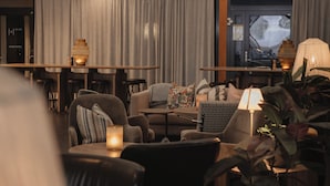 Ein gemütliches Wohnzimmer mit einer Couch, Sesseln, einem Couchtisch und einem Fernseher. Vorhänge, eine Lampe und eine Pflanze sind ebenfalls vorhanden.