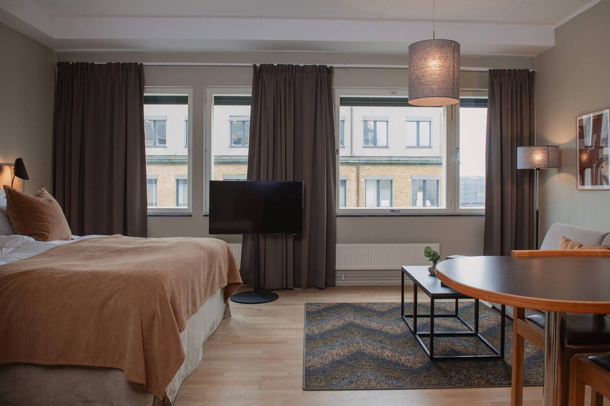 A hotel room with a bed, a TV, a dining table, and large windows with curtains.