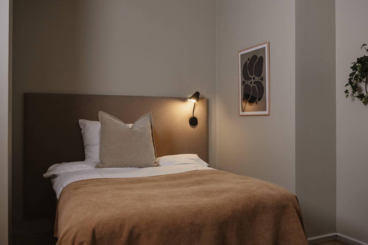 A neatly made bed with a brown headboard, a wall-mounted light, and a framed abstract artwork.