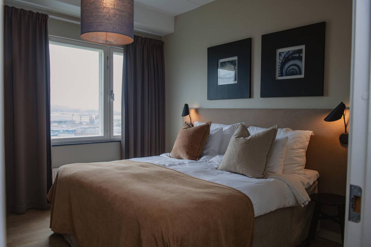 A bedroom with a large bed, a window with curtains, two wall-mounted lamps, and two framed pictures.