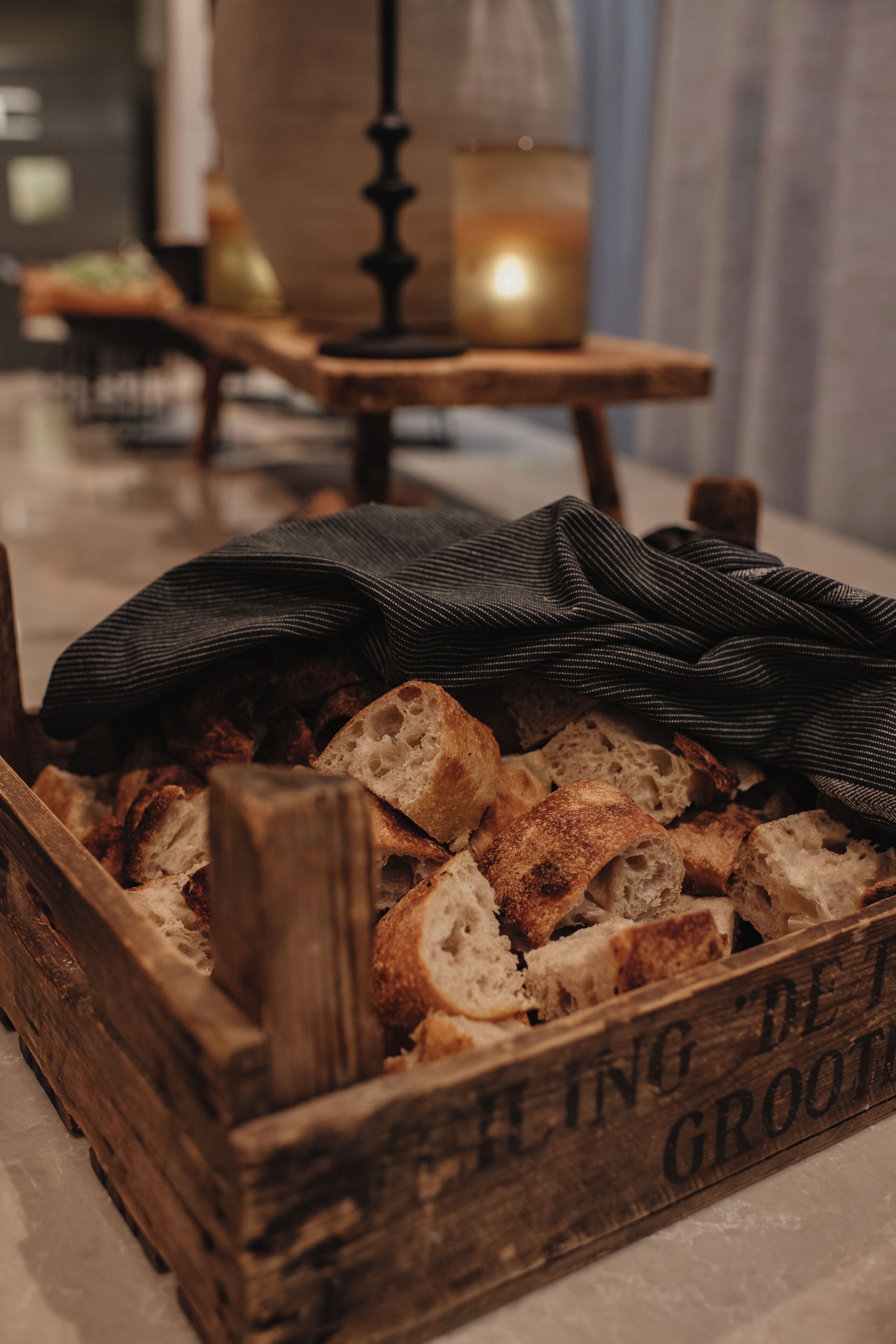 A wooden crate filled with sliced bread, a candle, and a cloth on a table in a room with a rustic decor.