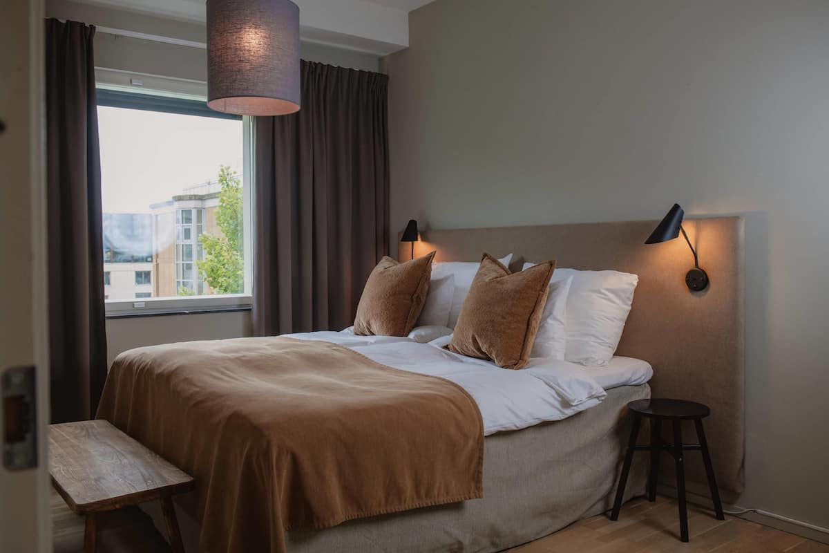 A hotel room with a bed, a bedside table, a lamp, and a view of buildings through a window.