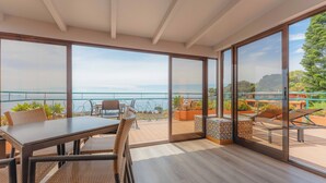 Un comedor moderno con una mesa grande y sillas, ventanas amplias con vistas al mar, y un balcón con mobiliario exterior.