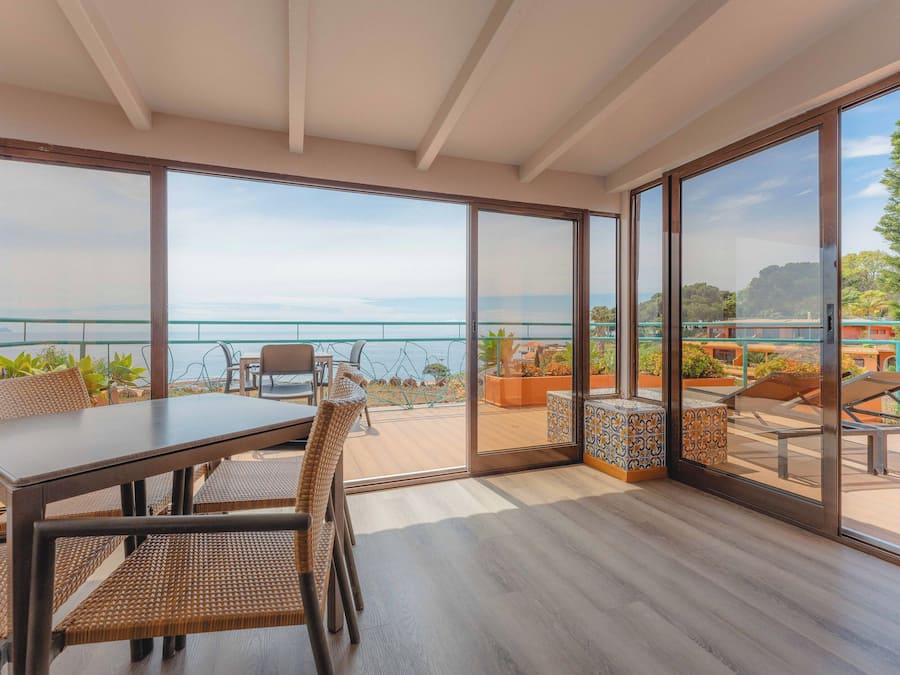 A modern dining area with a large table and chairs, large windows with a view of the sea, and a balcony with outdoor seating.