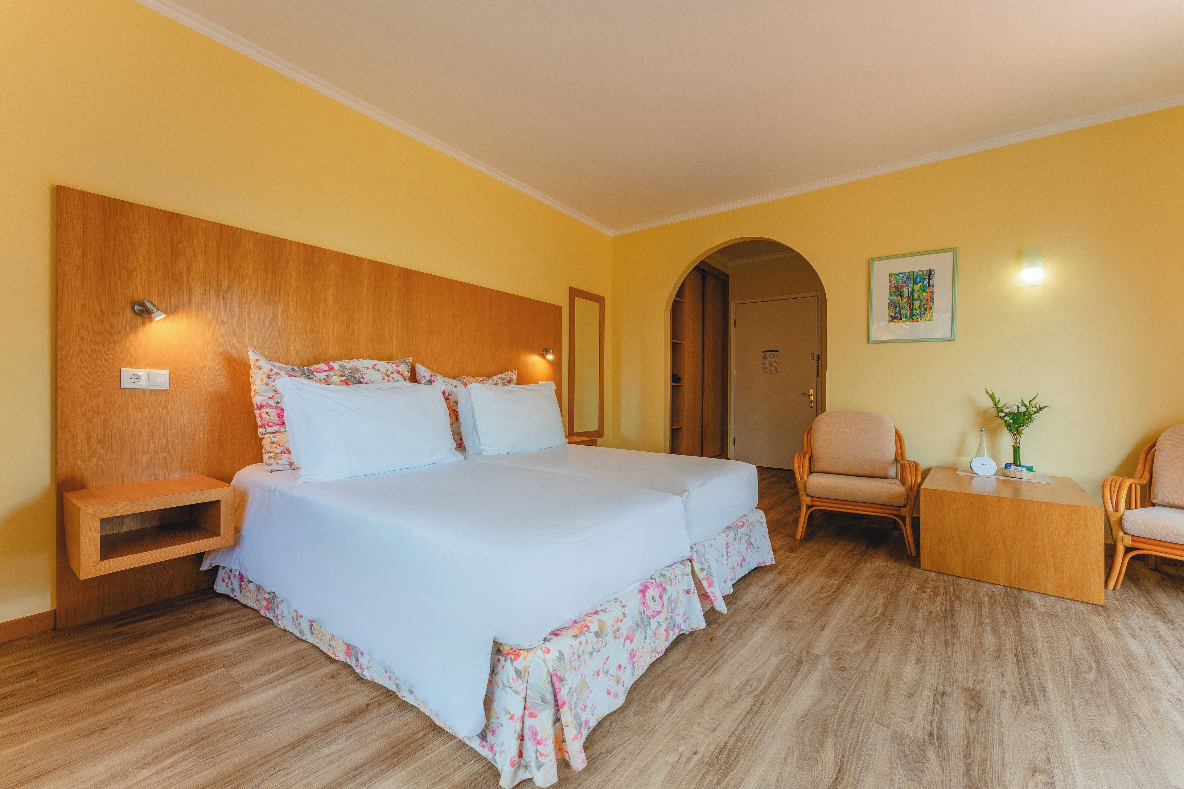 A hotel room with a large bed, wooden headboard, a nightstand, a desk with a chair, and a small table with a plant.