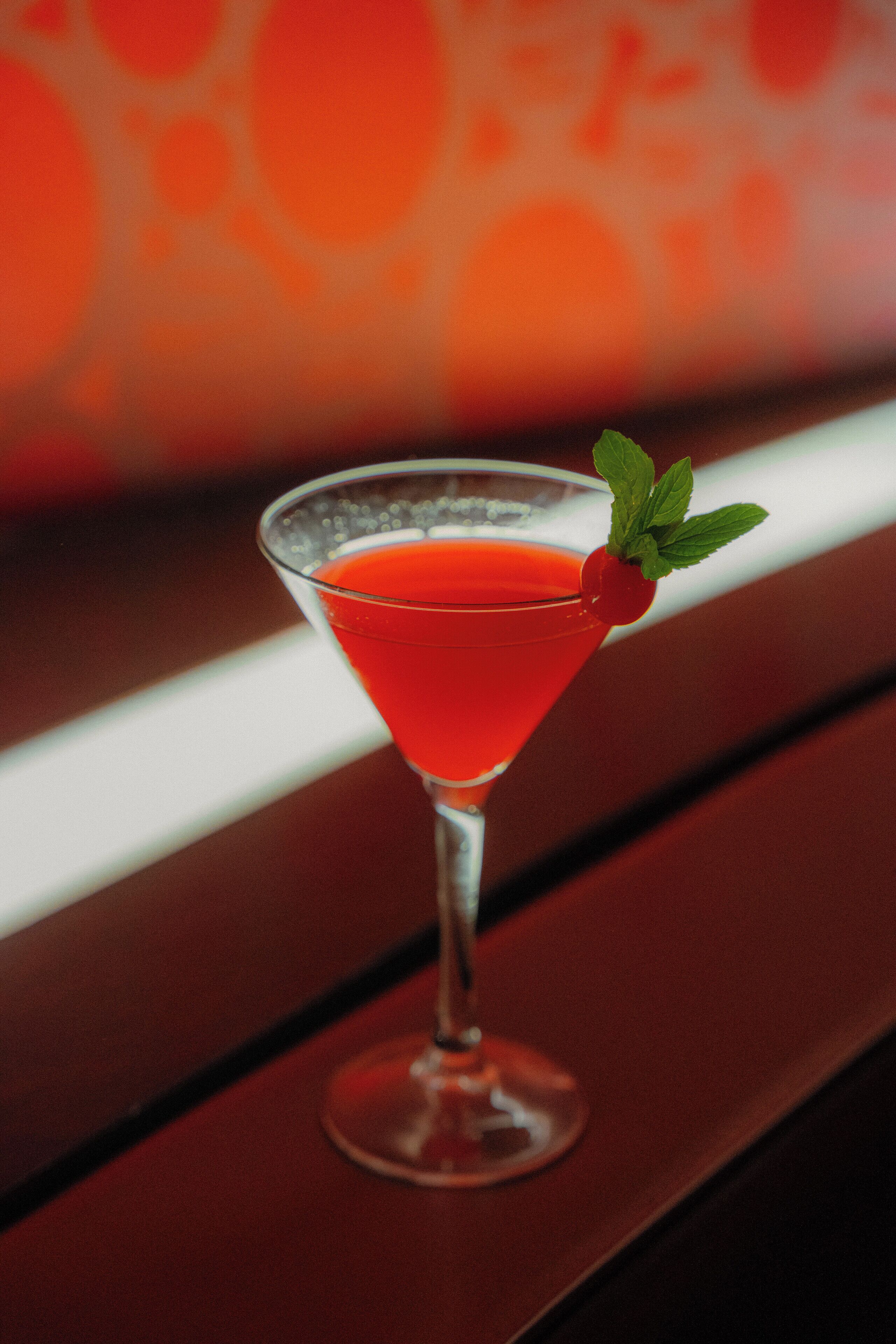 A martini glass with a red drink and a mint garnish on a wooden bar counter.