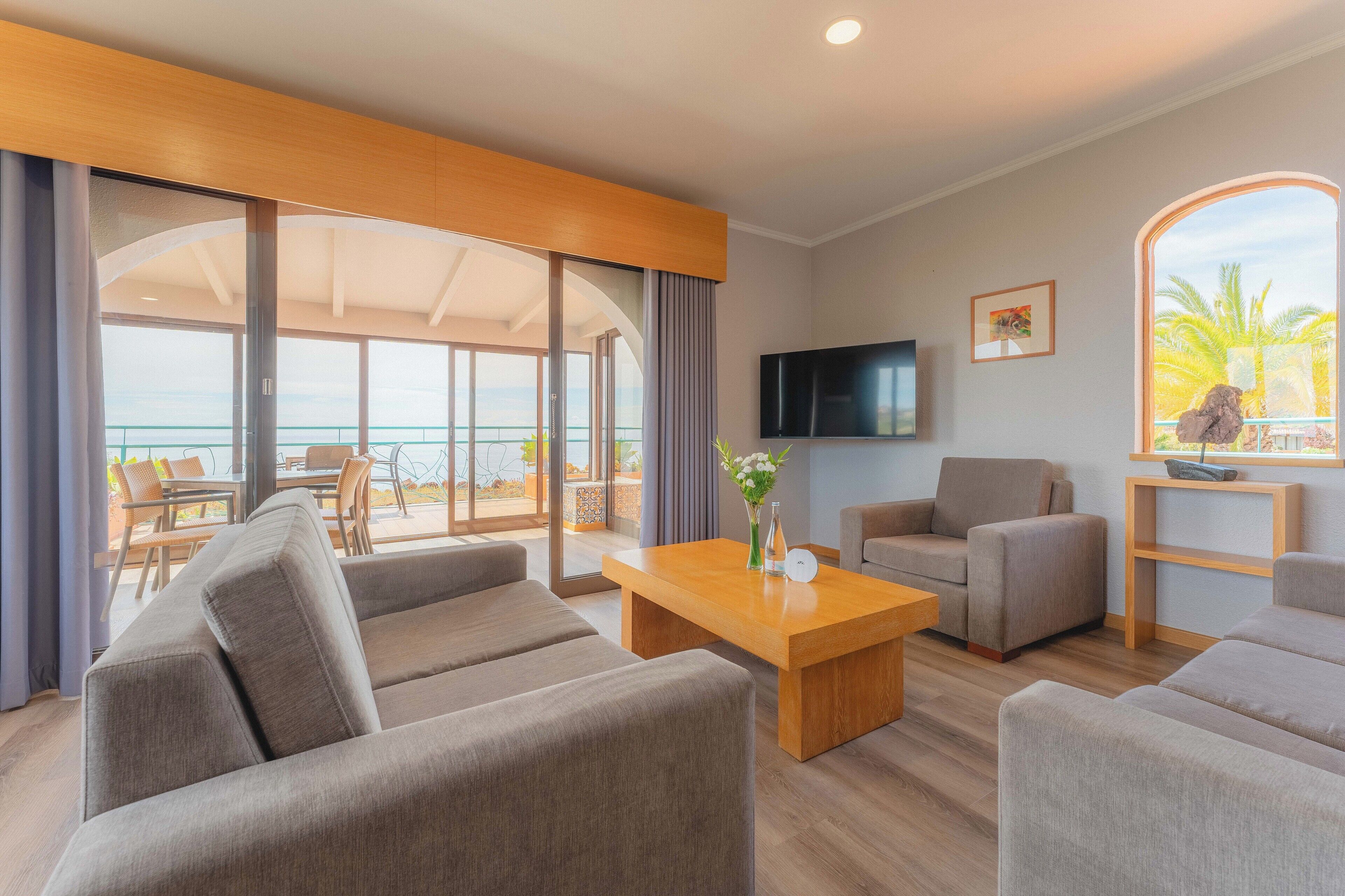 Un salón moderno con un gran ventanal corredera que da a un balcón con vistas al océano. Hay un televisor de pantalla plana montado en la pared y una mesa de centro de madera con un jarrón de flores.