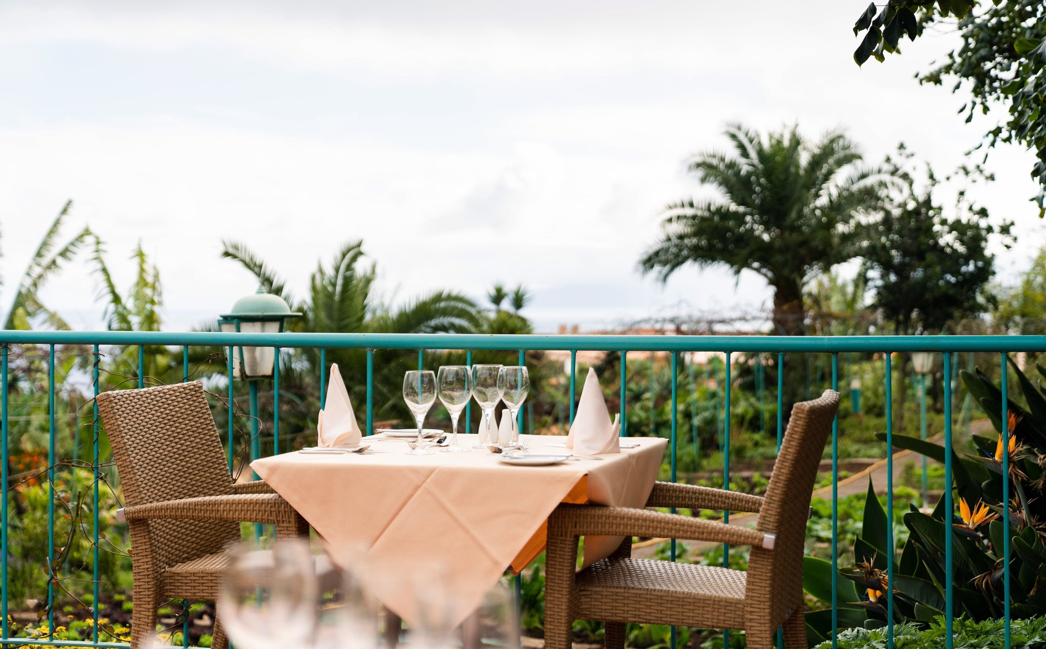 Un espace extérieur aménagé pour les repas, avec des chaises en osier et une table pour quatre personnes, donnant sur un jardin planté de palmiers et offrant une vue sur la mer.