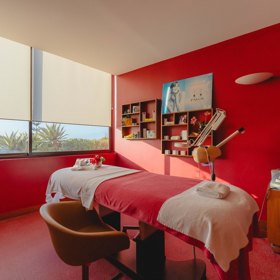 A room with a red wall, a bed, a chair, a small table, a shelf with books and a poster, and a window with a view of trees.