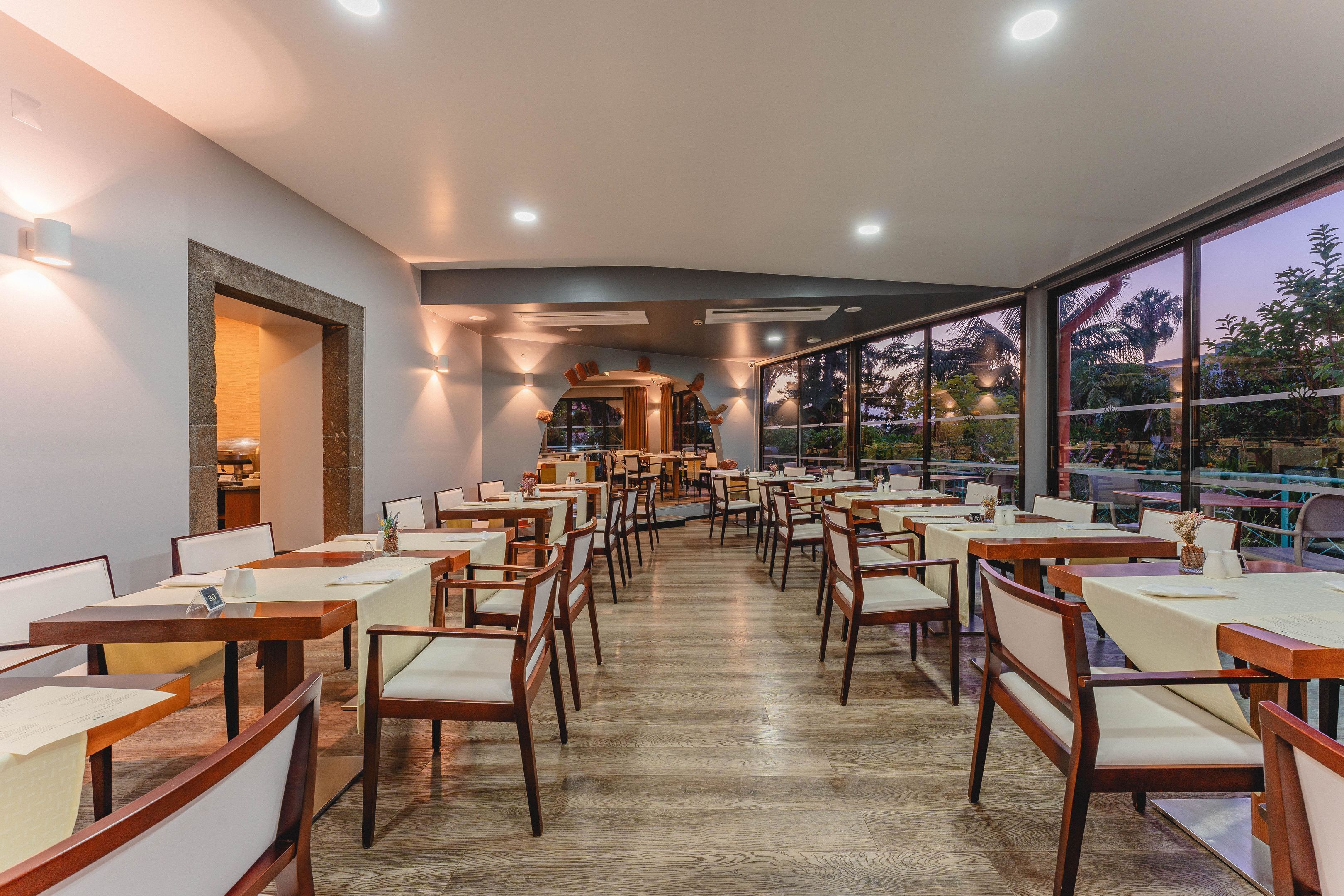 A spacious restaurant with wooden tables and chairs, a central bar area, and large windows offering a view of the outdoors.