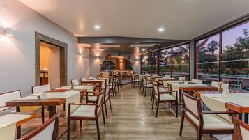 A spacious restaurant with wooden tables and chairs, a central bar area, and large windows offering a view of the outdoors.