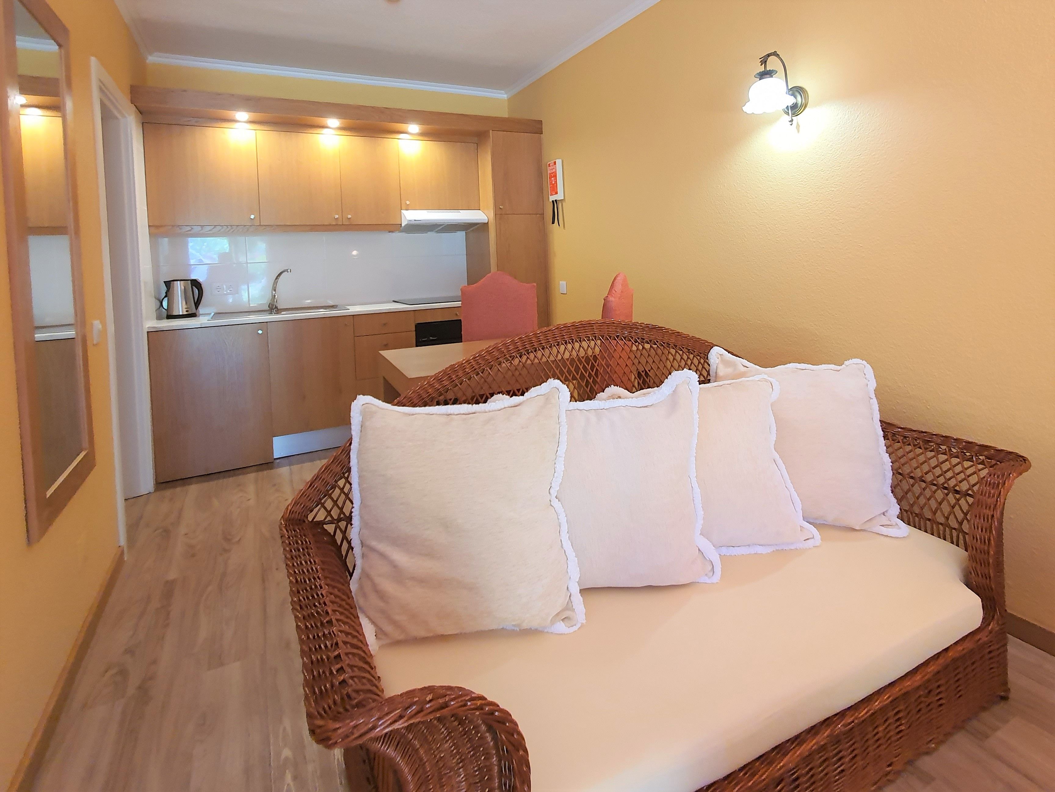 A room with a wicker sofa, a kitchen area in the background, and a wall-mounted light fixture.