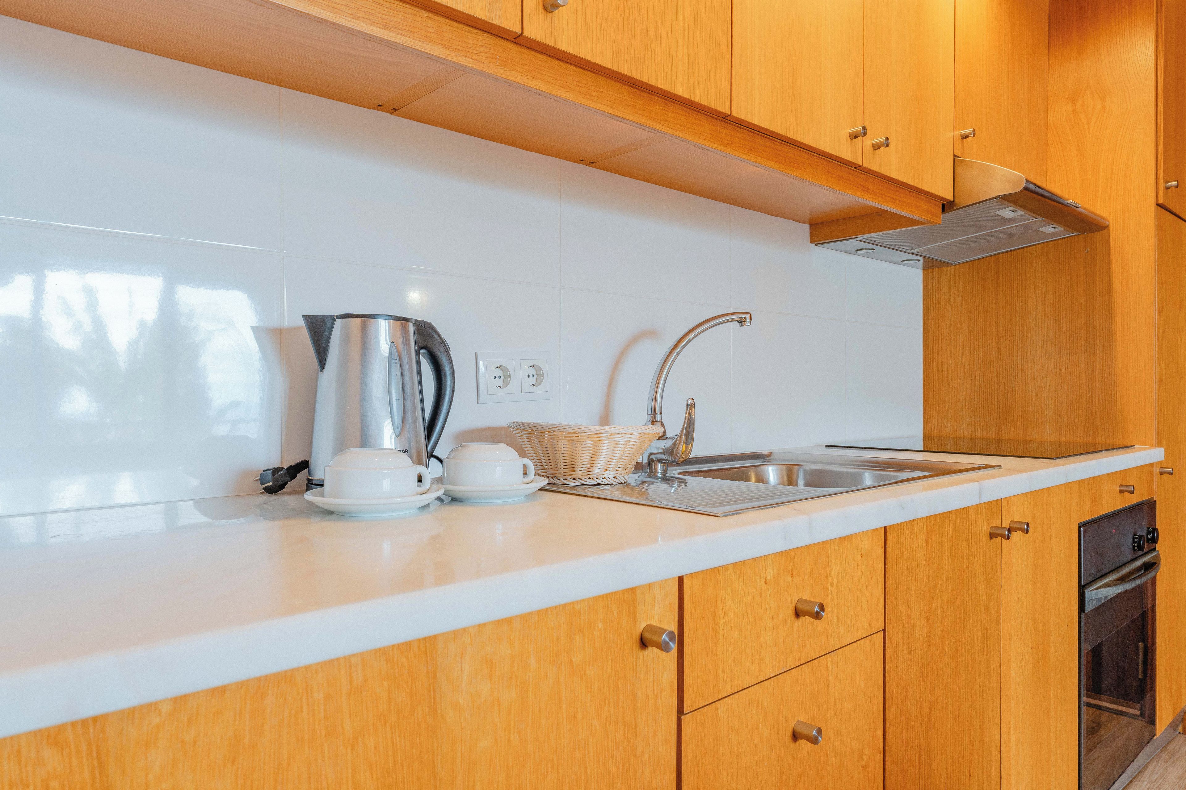 A modern kitchen with wooden cabinets, a white countertop, a stainless steel kettle, a coffee maker, and a sink.