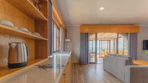 Una habitación de hotel moderna con una zona de cocina, un sofá gris y un amplio ventanal corredero que da a un balcón con vistas.