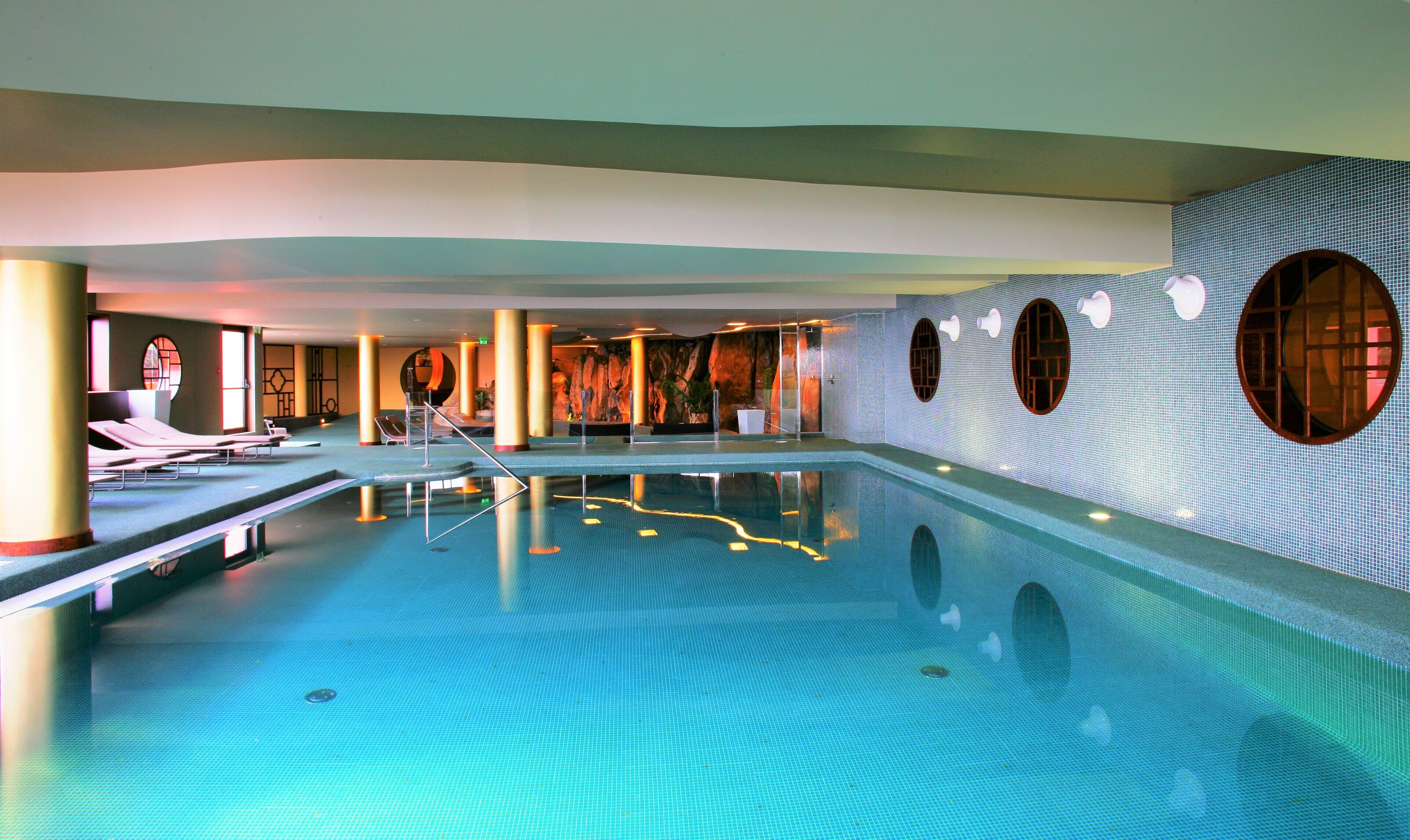 Una amplia piscina interior con superficie de agua azul cristalina, rodeada de sillones de descanso y un diseño moderno con ventanales circulares y azulejos decorativos.