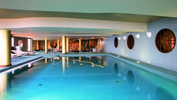 Una amplia piscina interior con superficie de agua azul cristalina, rodeada de sillones de descanso y un diseño moderno con ventanales circulares y azulejos decorativos.