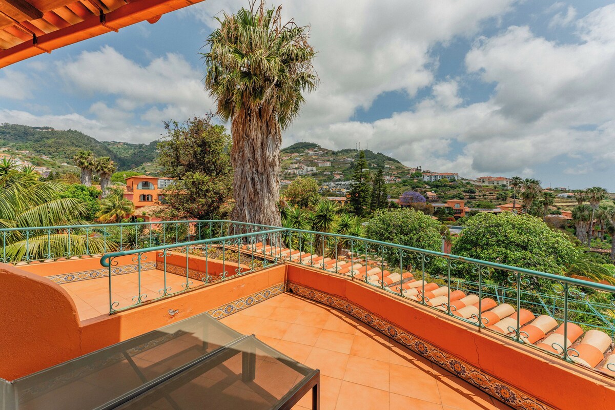 A terrace with orange walls and a glass table, overlooking a hillside with buildings and greenery.