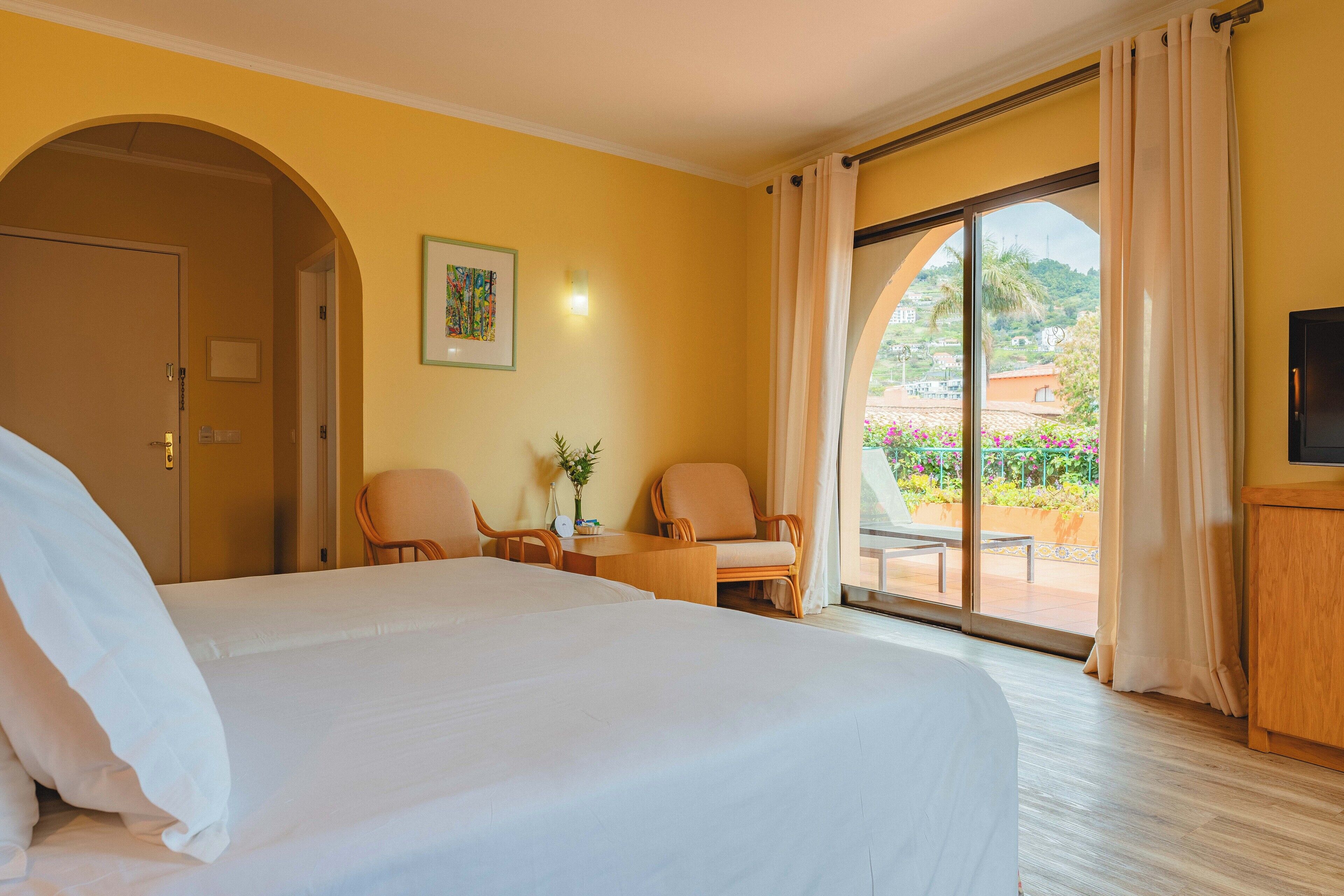 A hotel room with a large bed, two armchairs, a small table with a vase, and a view of a garden through a sliding glass door.