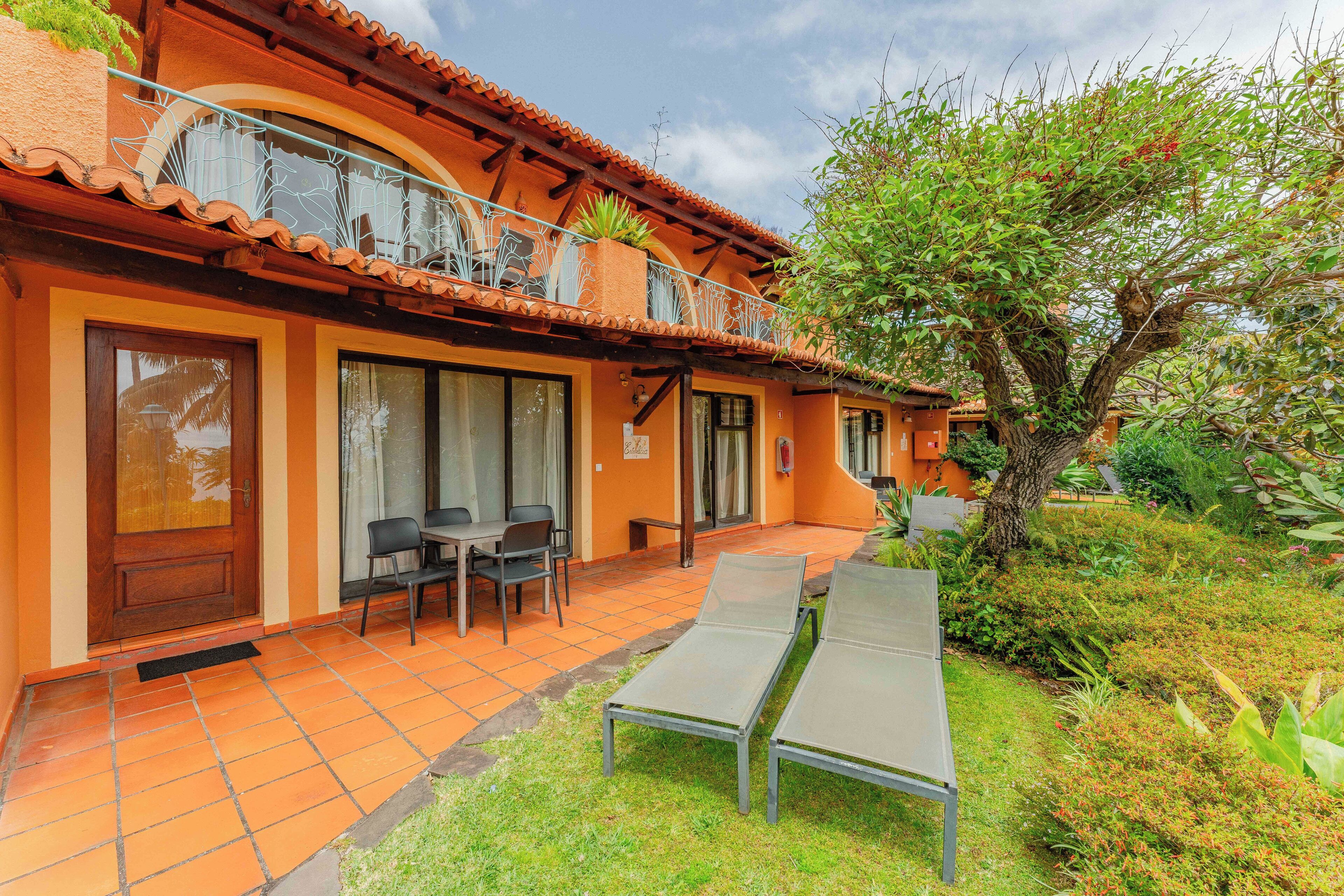 Un edificio con un patio empedrado, un comedor con sillas y un pequeño jardín con un árbol y plantas.