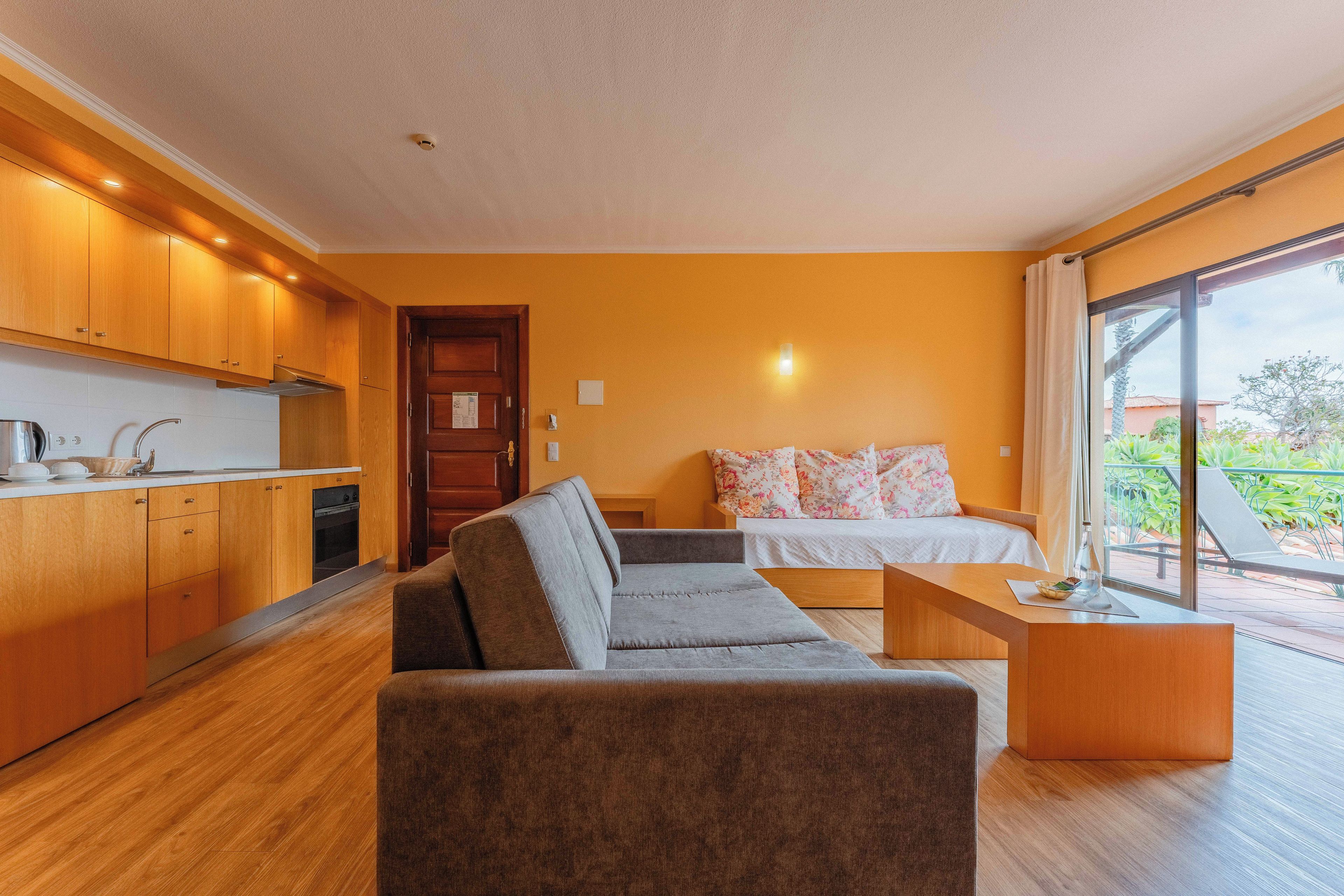A modern living room with a sofa, a small table, and a bed with floral bedding. There is a kitchen area with wooden cabinets and a window overlooking a balcony.