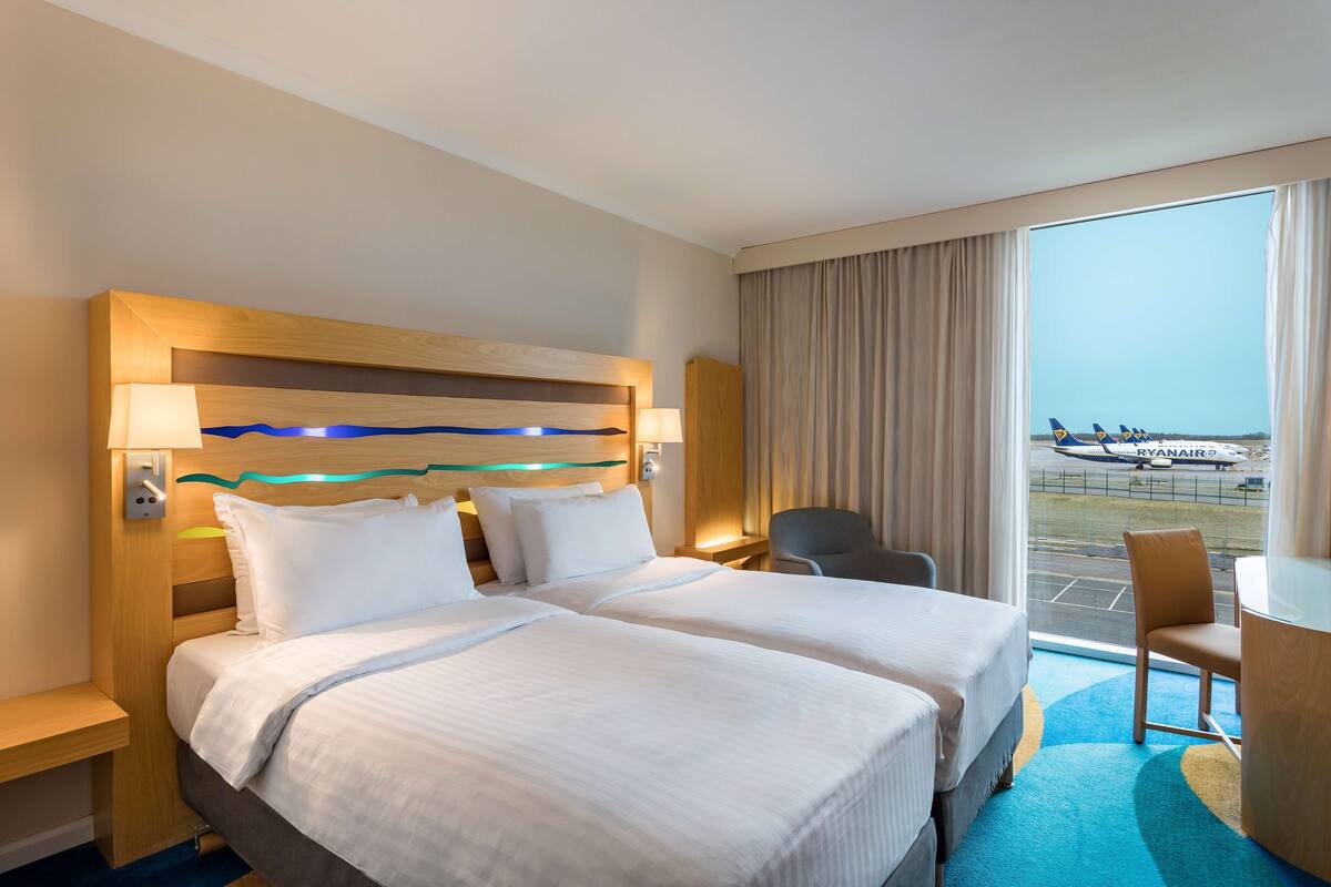 A hotel room with two beds, a desk, and a view of an airport.