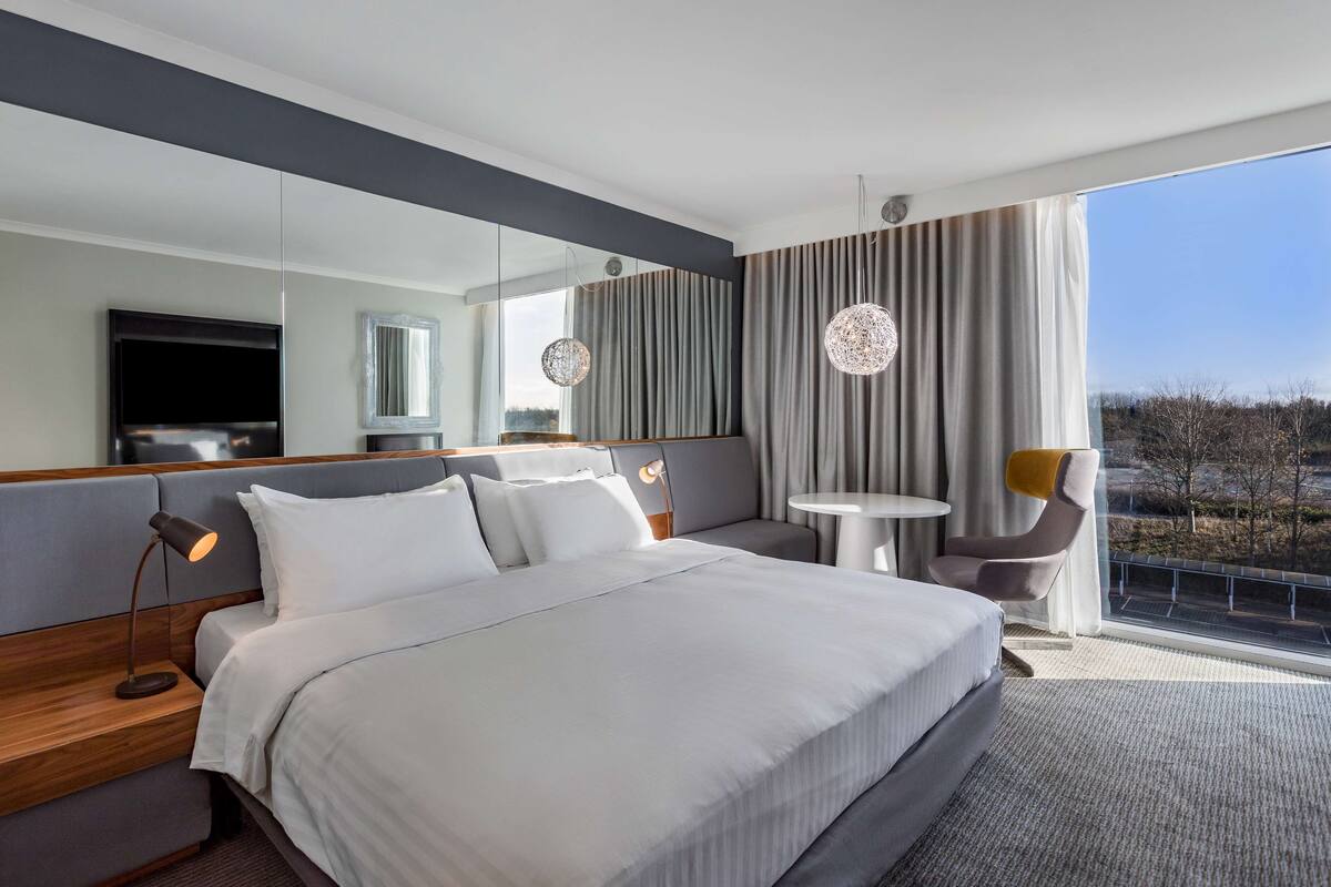 A modern hotel room with a large bed, a sitting area with a table and chairs, a TV, and a large window with a view.