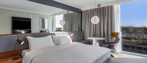 Una habitación de hotel moderna con una cama grande, una zona de estar con una mesa y sillas, un televisor y un ventanal con vistas.