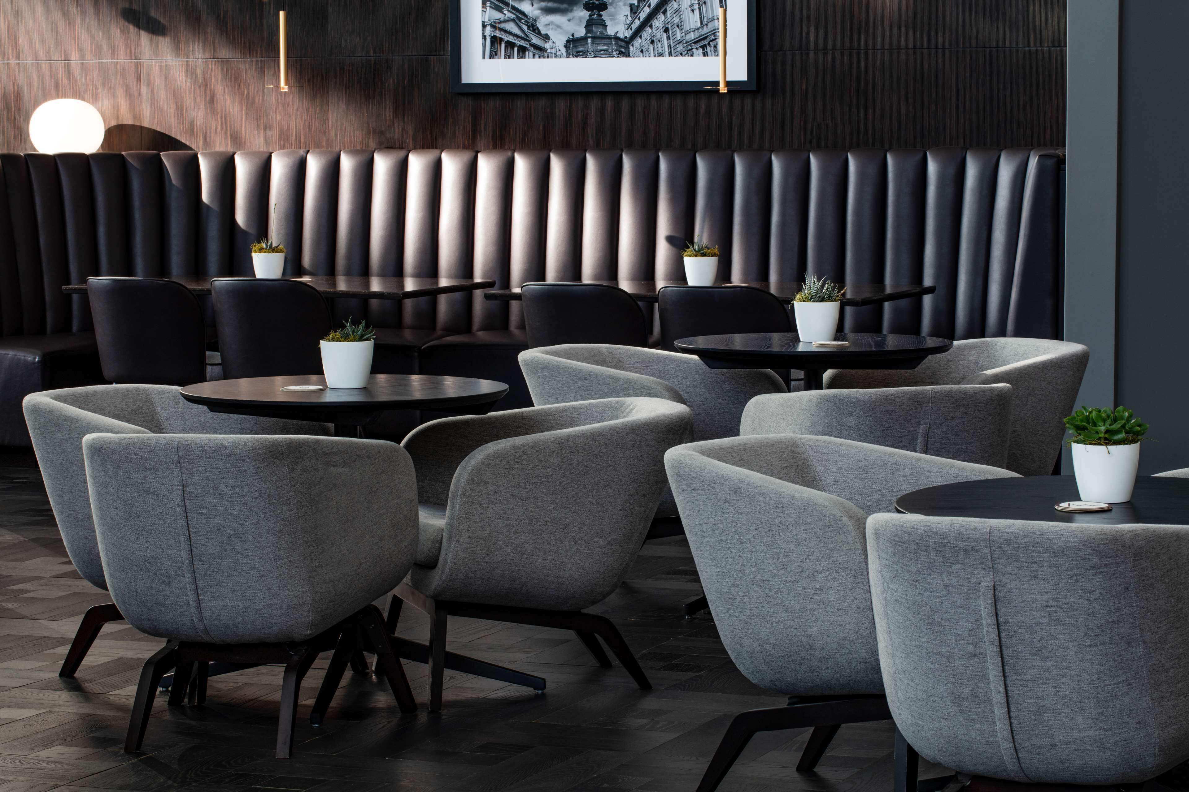A modern restaurant with grey upholstered chairs and tables, a dark wood panel wall, and a framed picture above the seating area.